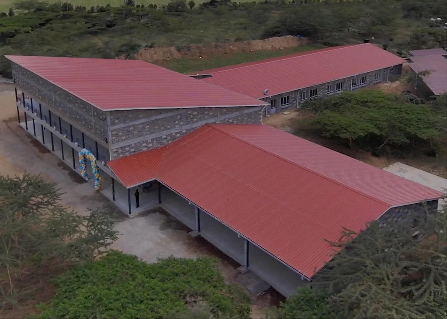 Aerial view of a school building with a red roof, stone walls, surrounding trees, and balloons at the entrance, likely for a celebration.