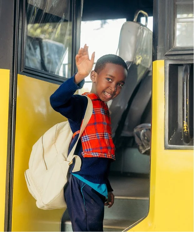 A young boy with a backpack standing in the doorway of a yellow school bus, smiling and waving.