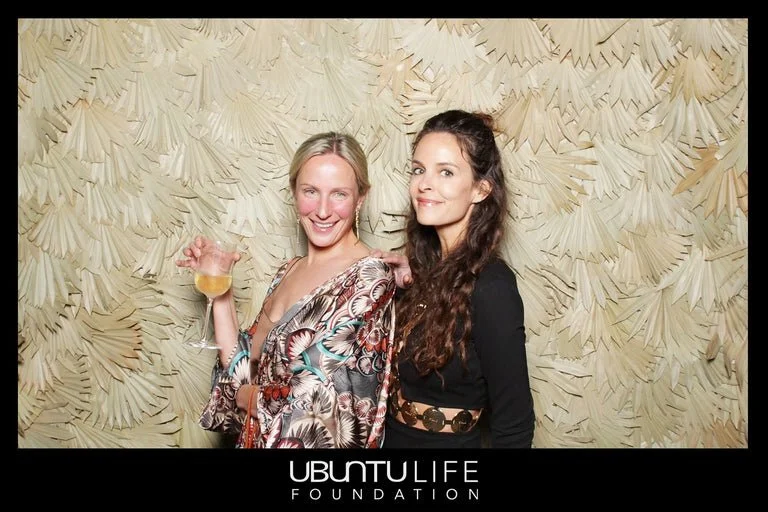 Two women smiling and posing for a photo at an event, with a decorative cream-colored textured background. One woman holds a wine glass. There is an 'Ubuntu Life Foundation' logo at the bottom.