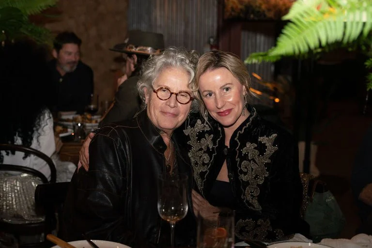 Two women sitting close together at a restaurant, smiling for the camera. The older woman has gray hair and glasses, and the younger woman has blonde hair and is wearing a black and gold embroidered jacket. There are drinks on the table and other peo