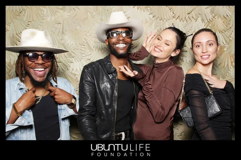 Four diverse young adults smiling and posing together in front of a textured background, with two men wearing wide-brimmed hats and glasses, and two women with stylish outfits. They appear to be at a social event or gathering, with a logo for the Ubu