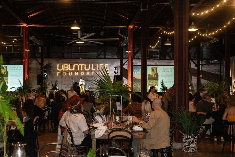 A crowded indoor event space with people seated at tables, decorated with large potted plants, string lights, and a neon sign that says 'Ubuntulu Foundation.'