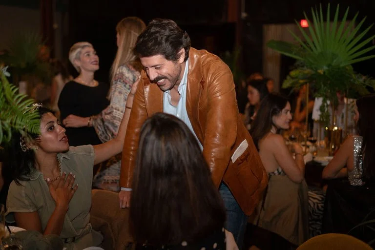 Man in brown leather jacket smiling and talking to seated woman in green dress at a social gathering or party.