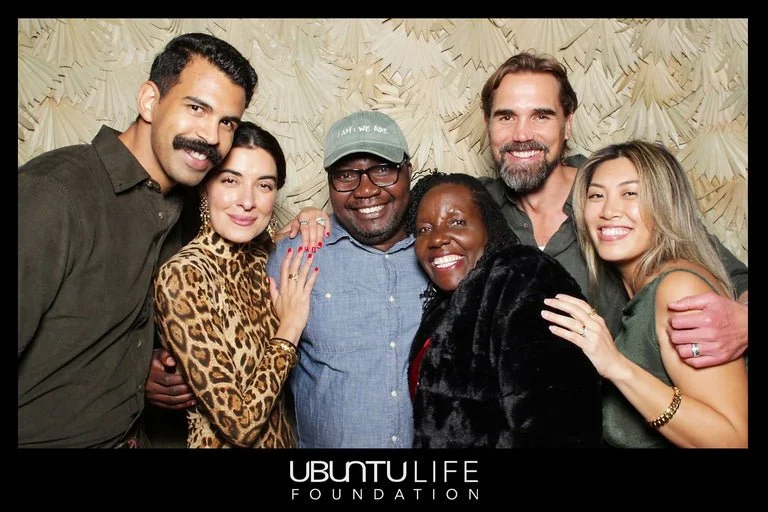 Group of six diverse people smiling and posing for a photo together, with a beige textured backdrop and the Ubuntu Life Foundation logo at the bottom.