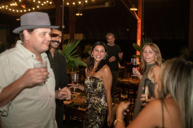 Group of people at a social gathering or party, some holding drinks, with warm lighting and decorative plants in the background.