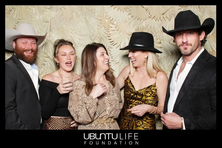 Group of five people dressed in stylish outfits and cowboy hats, smiling and chatting at a social event, with a backdrop featuring a textured pattern and the 'Ubuntu Life Foundation' logo.
