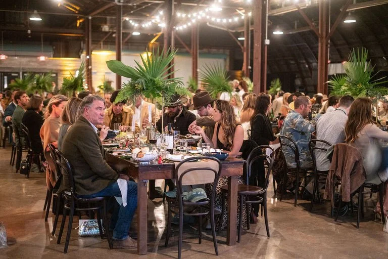 People dining at a long table decorated with large green tropical plants, under string lights in a rustic indoor setting.