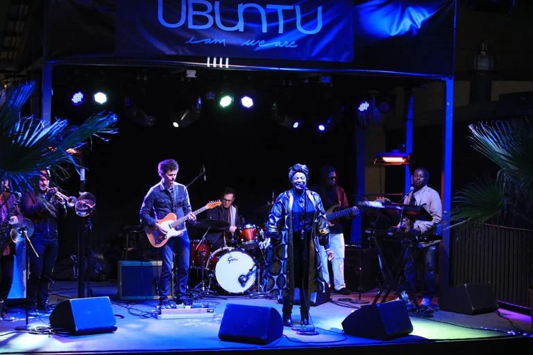 A live band performing on stage at an event with a blue banner that says 'UBUNTU'. The band includes a woman singer, a guitarist, a drummer, and keyboard players, with stage lights illuminating the scene.