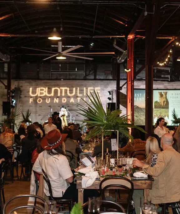A group of people dining at tables in a dimly lit venue with a sign that reads 'UBUNTULIFE FOUNDATION' on the wall, large plants on the tables, and projected images on screens in the background.