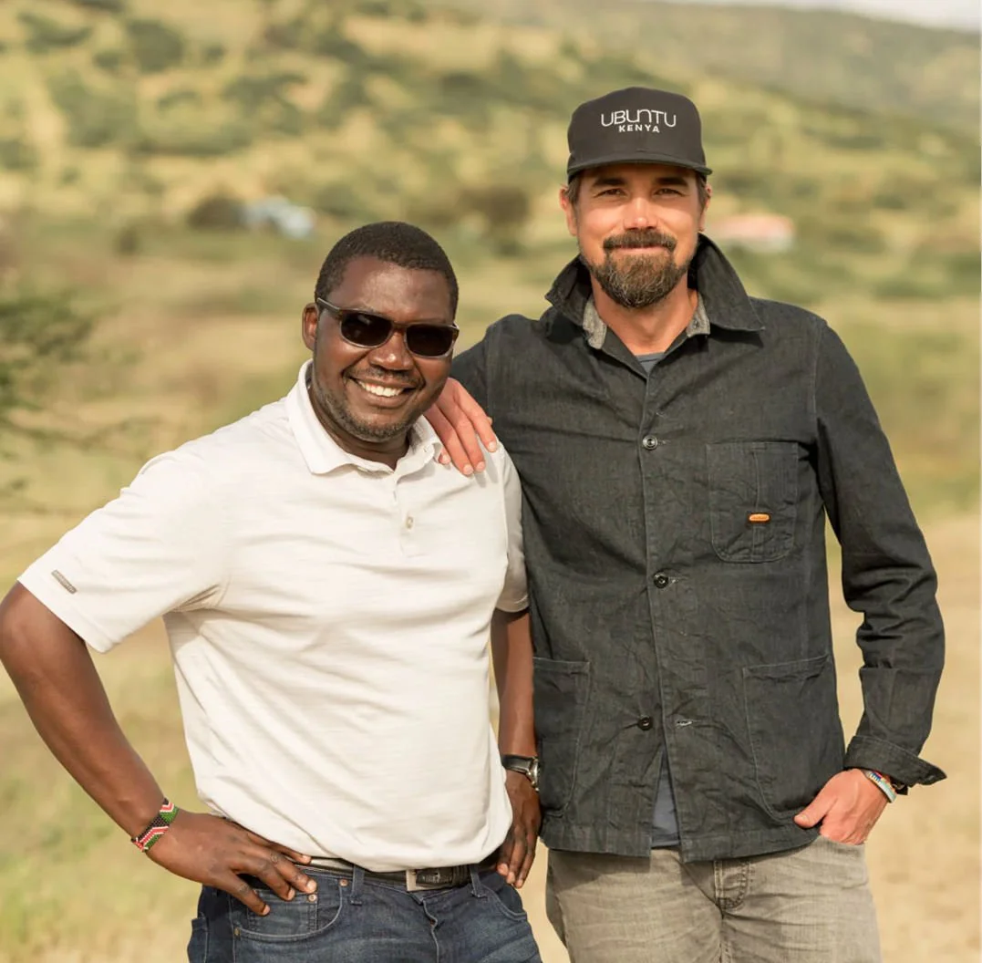 Two men standing outdoors with a blurred natural background. The man on the left is wearing a white polo shirt, dark sunglasses, and a wristband, smiling with his hand on his hip. The man on the right is wearing a dark button-up jacket, a cap that says UBTU Kenya, and has a beard, smiling with one hand in his pocket and the other resting on the other man's shoulder.