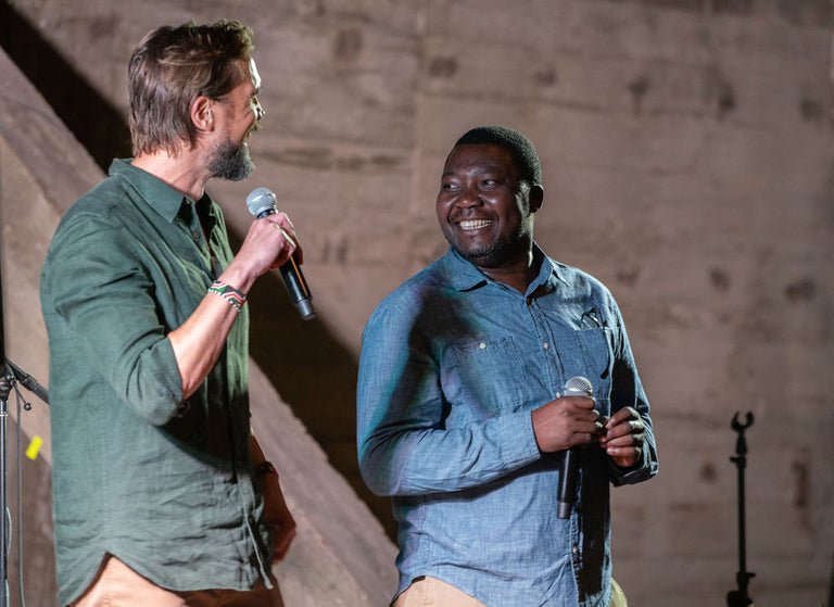 Two men on stage, one holding a microphone, smiling at each other, in front of a wooden background.
