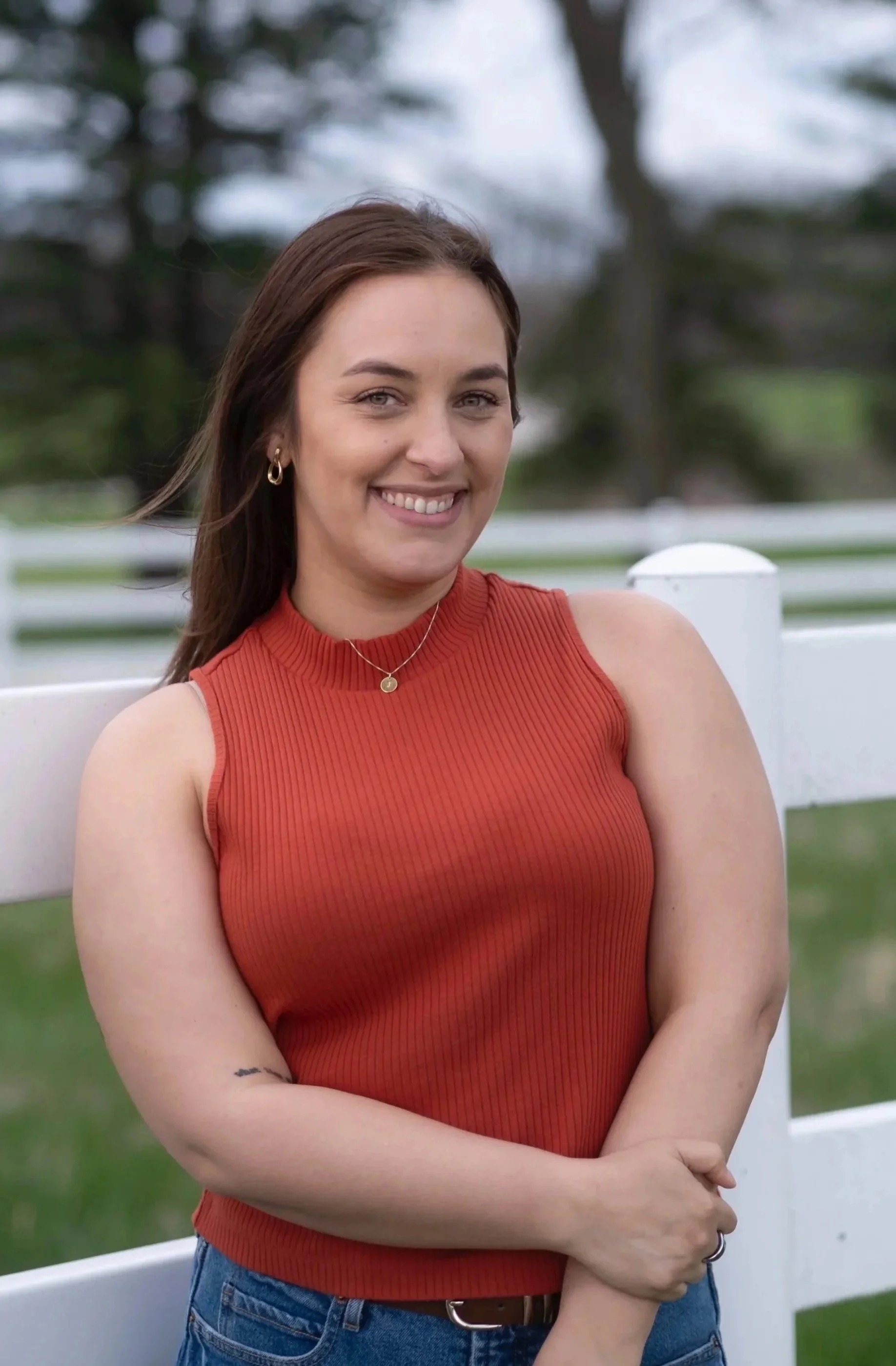 A young woman with long brown hair smiling and posing outdoors, wearing a sleeveless red top, gold jewelry, and blue jeans, leaning against a white fence with a blurred background of trees and grass.