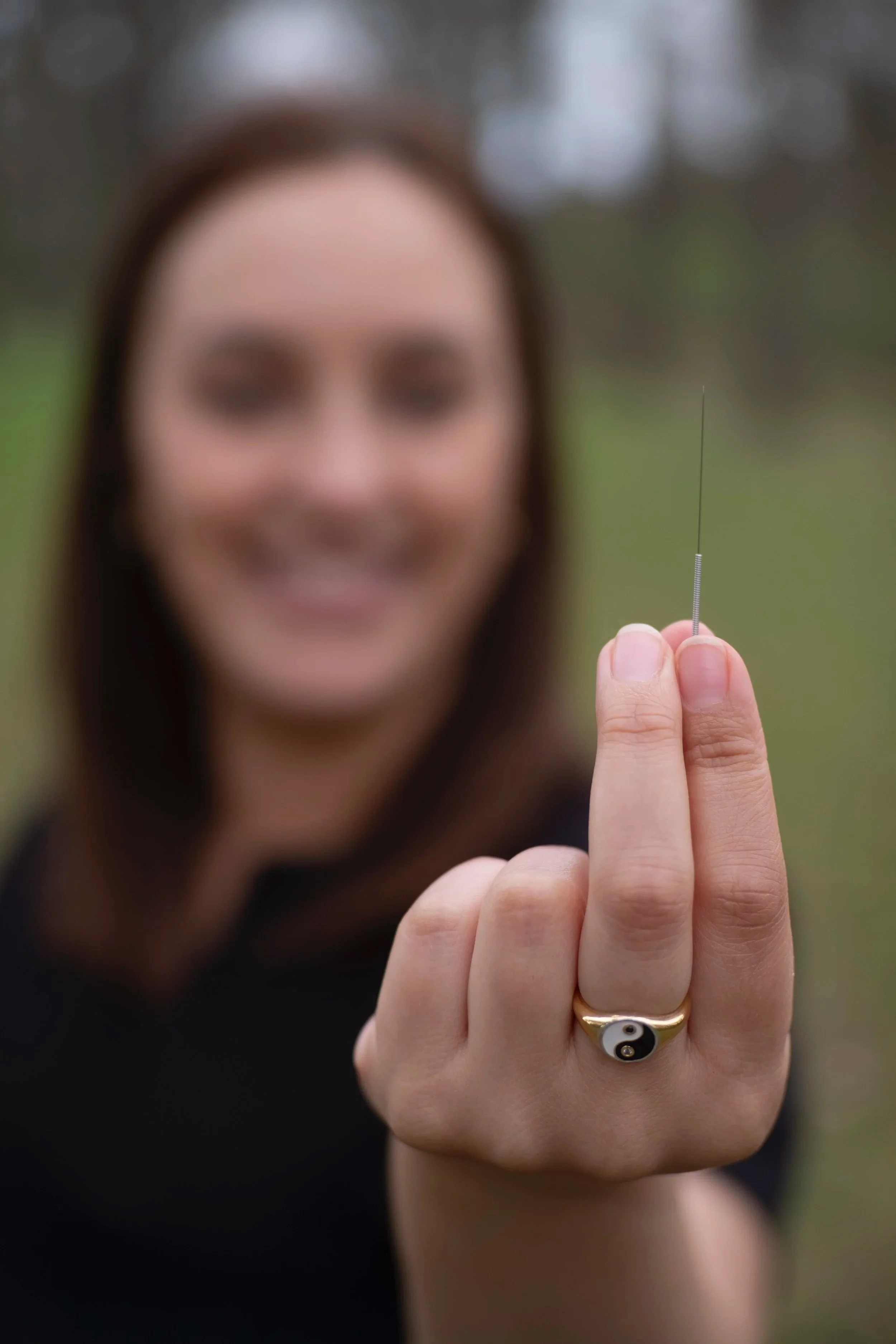 A woman with a Yin-Yang ring on her finger holding a thin acupuncture needle with her hand extended towards the camera, her face blurred in the background.