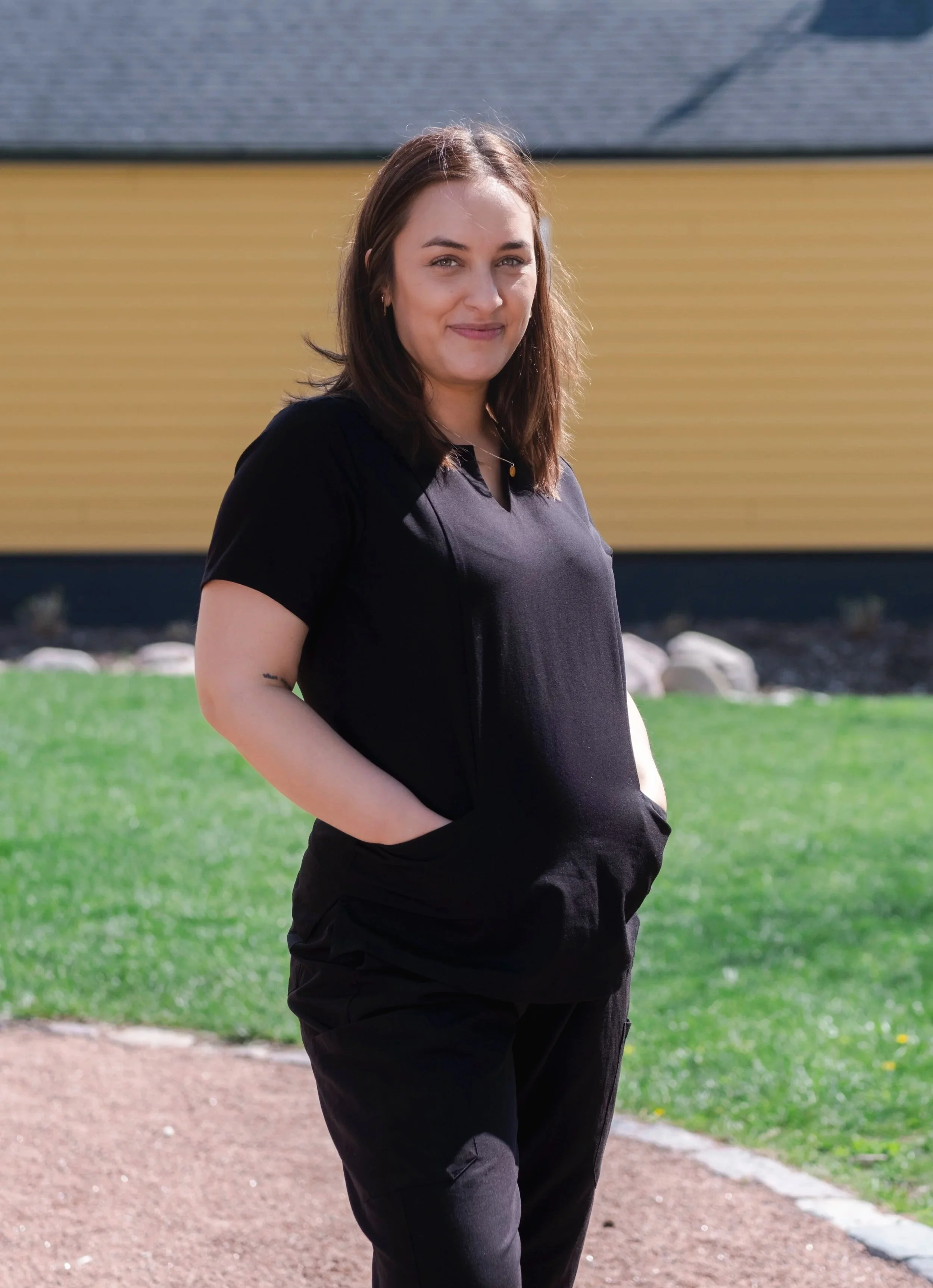 A young woman with shoulder-length brown hair and light skin standing outdoors on a sunny day. She is wearing a black t-shirt and black pants, with her hands in her pockets, smiling at the camera. The background includes a yellow building, green grass, and some rocks.