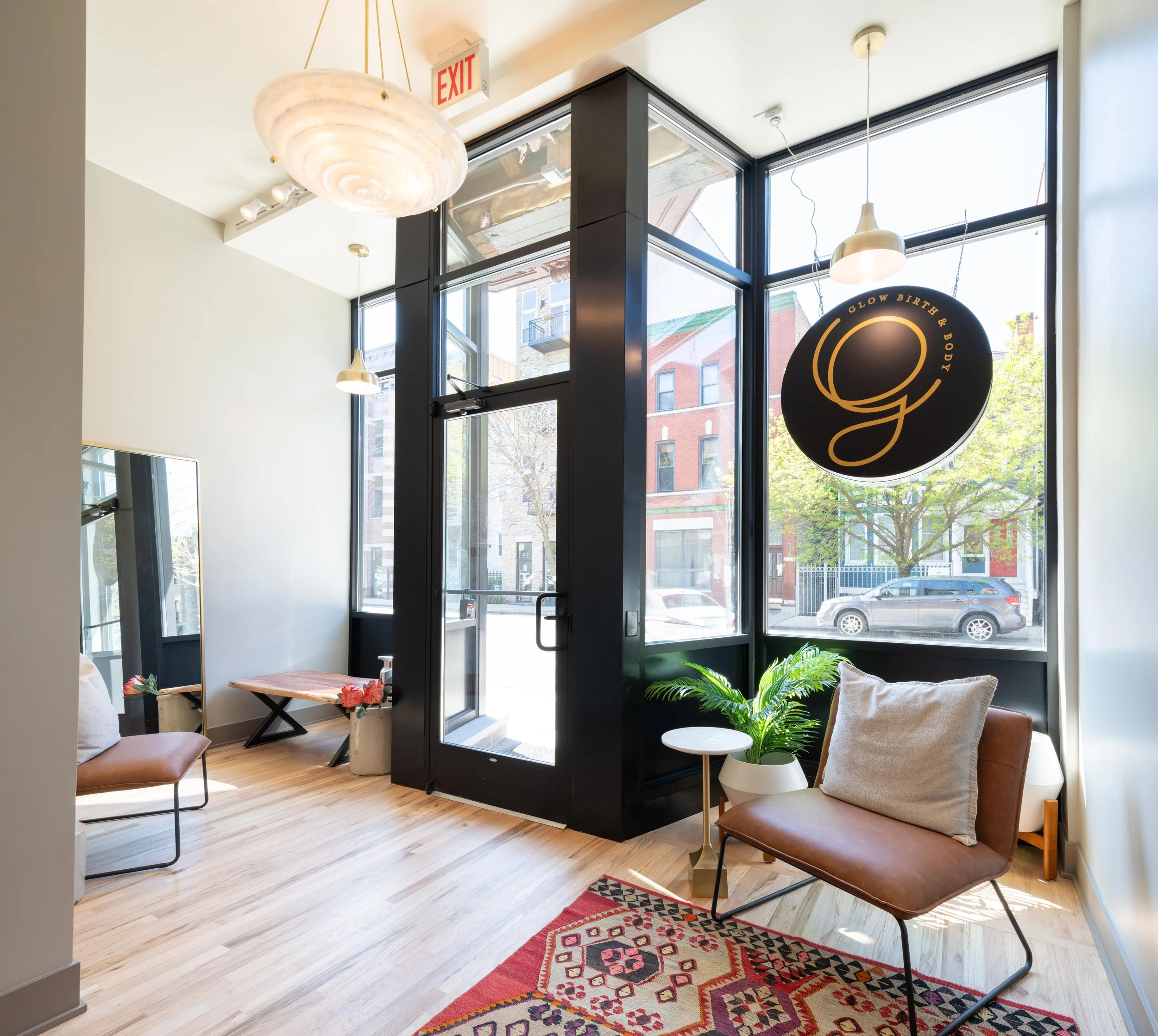 Interior of a modern boutique with large front windows, wooden floors, a leather chair with a pillow, a white side table with a plant, a colorful rug, and a circular sign hanging from the ceiling reading "Glow, Birth & Body."