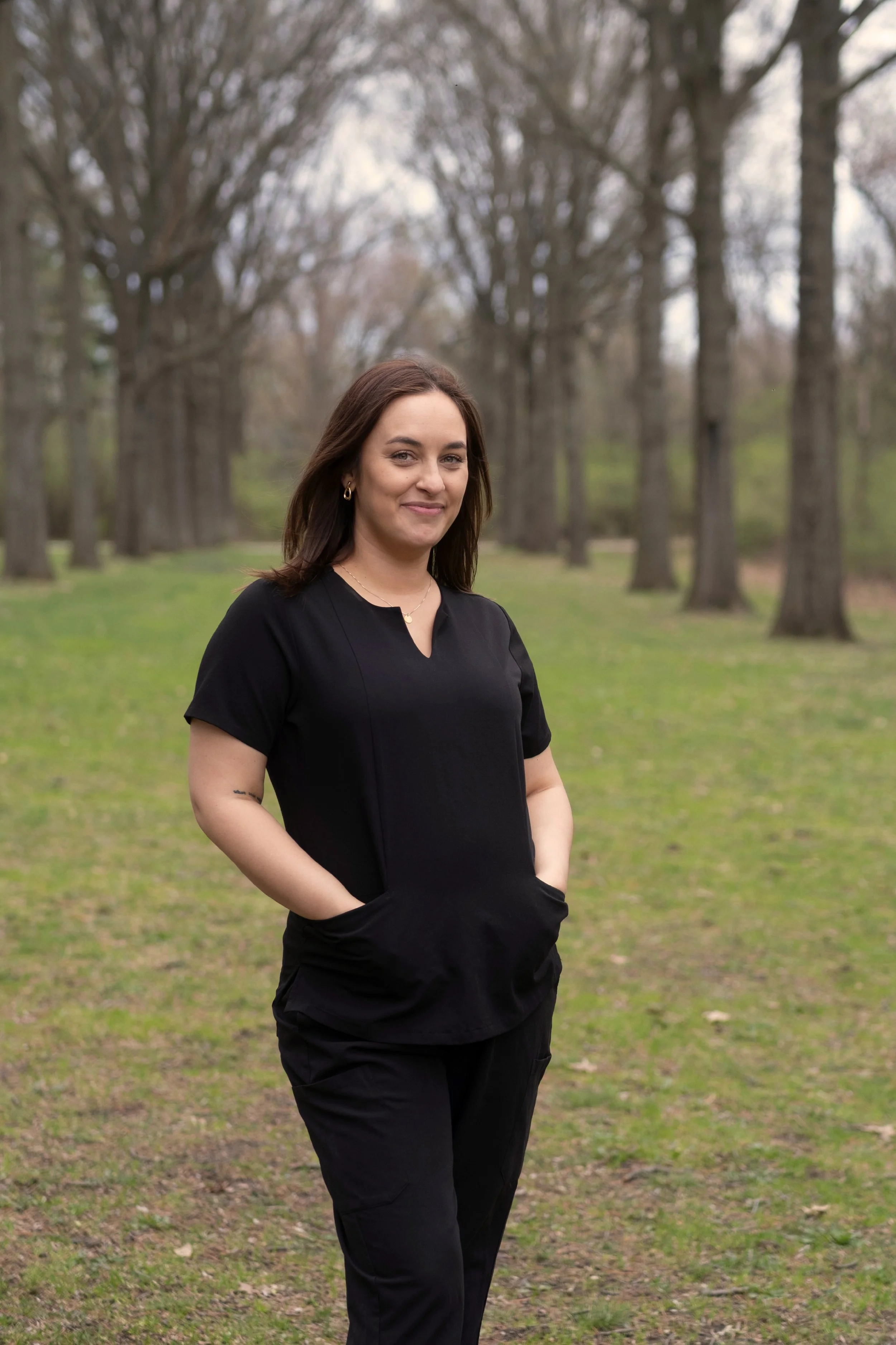 A woman standing in a park with trees in the background, wearing black clothing, with hands in her pockets and smiling.