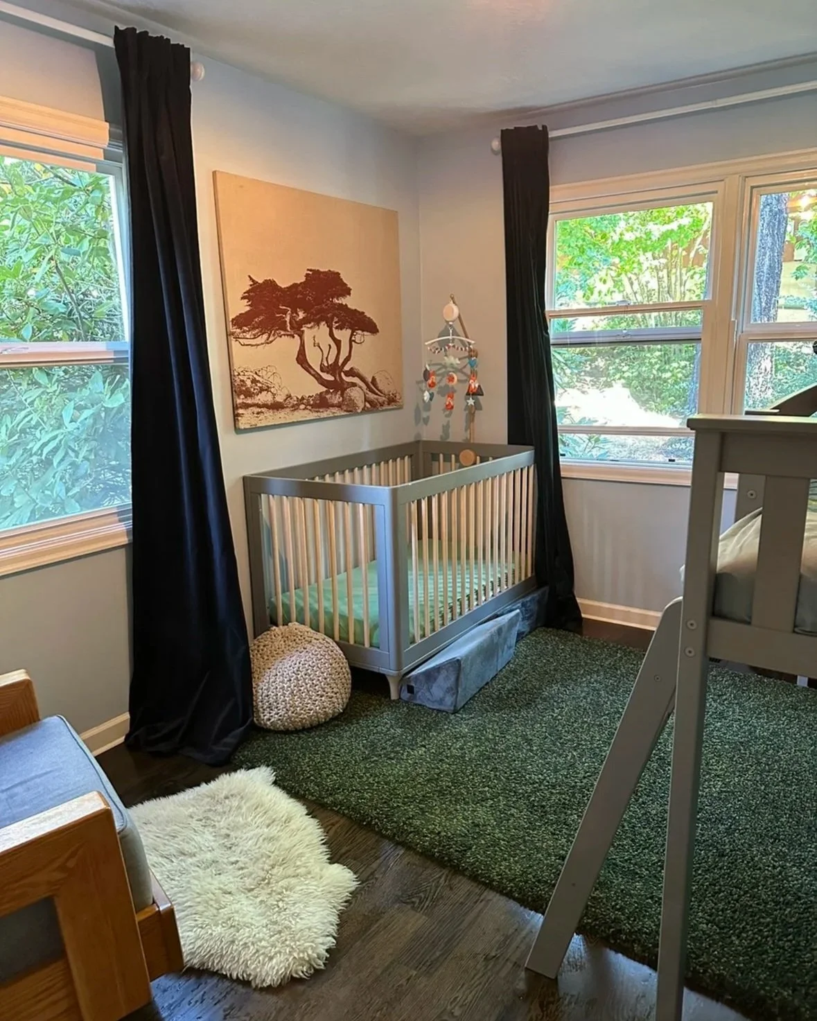 A nursery with two large windows, black curtains, a framed tree artwork on the wall, a crib with a green mattress, a white fluffy rug, a textured round pouf, and a wall hanging mobile with animals.