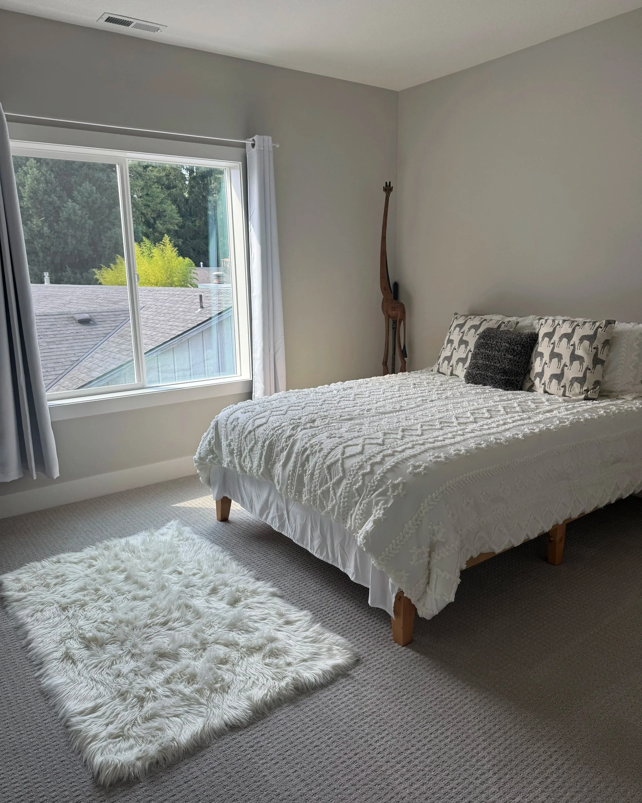 A bedroom with a large window, a bed with white textured bedding, a black and white pillow, a small furry rug on beige carpet, and a wooden giraffe sculpture in the corner.