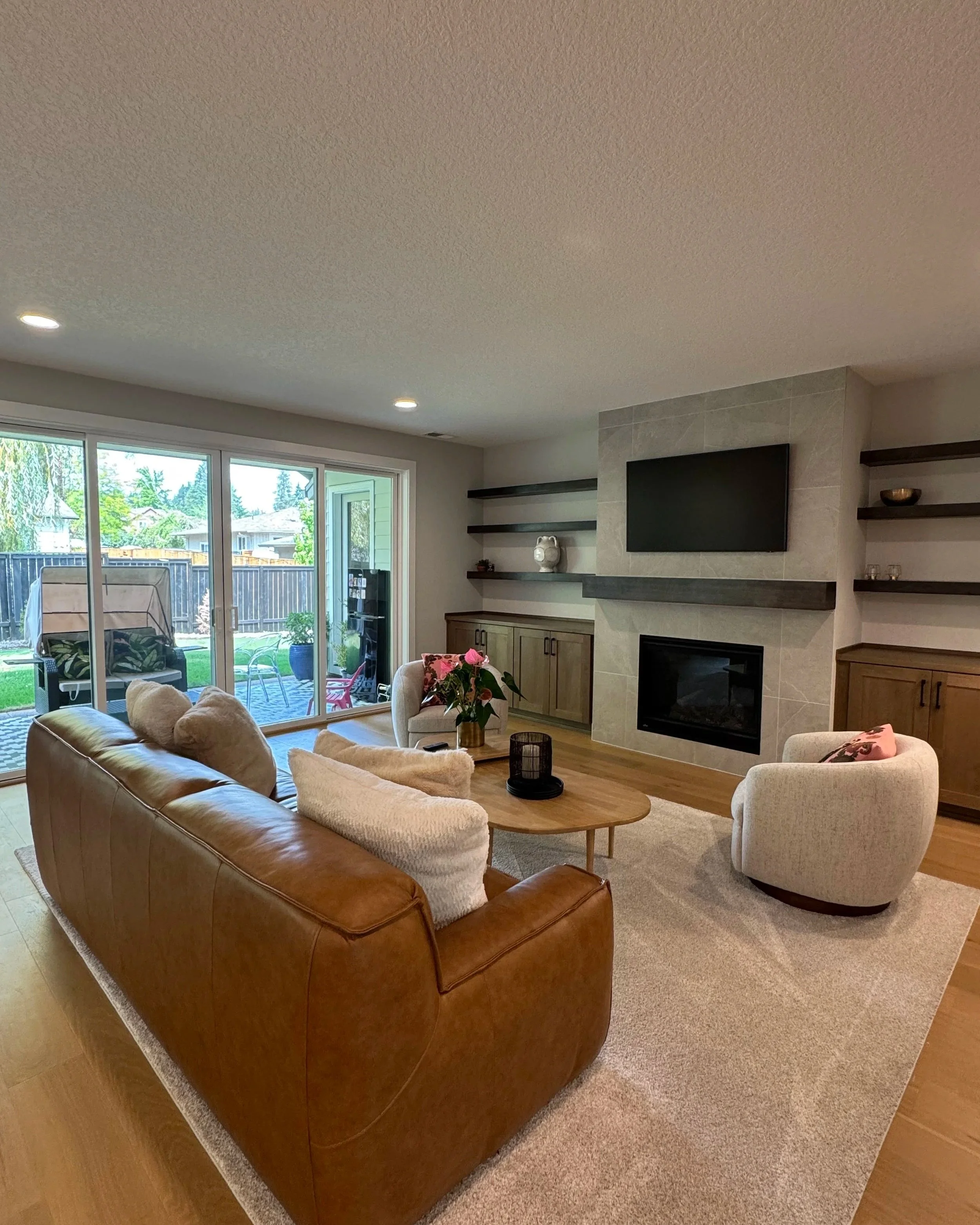 Living room with brown leather sofa, white armchairs, a wooden coffee table, a fireplace, shelves, and a large sliding glass door leading to an outdoor space.