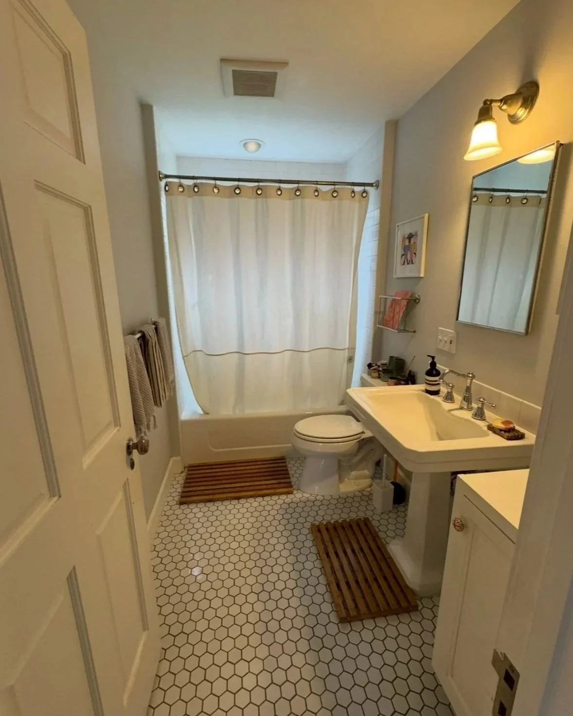 Small bathroom with white hexagon floor tiles, a white bathtub with a cream curtain, a white toilet, a white sink with a mirror above, and wooden bath mats on the floor.