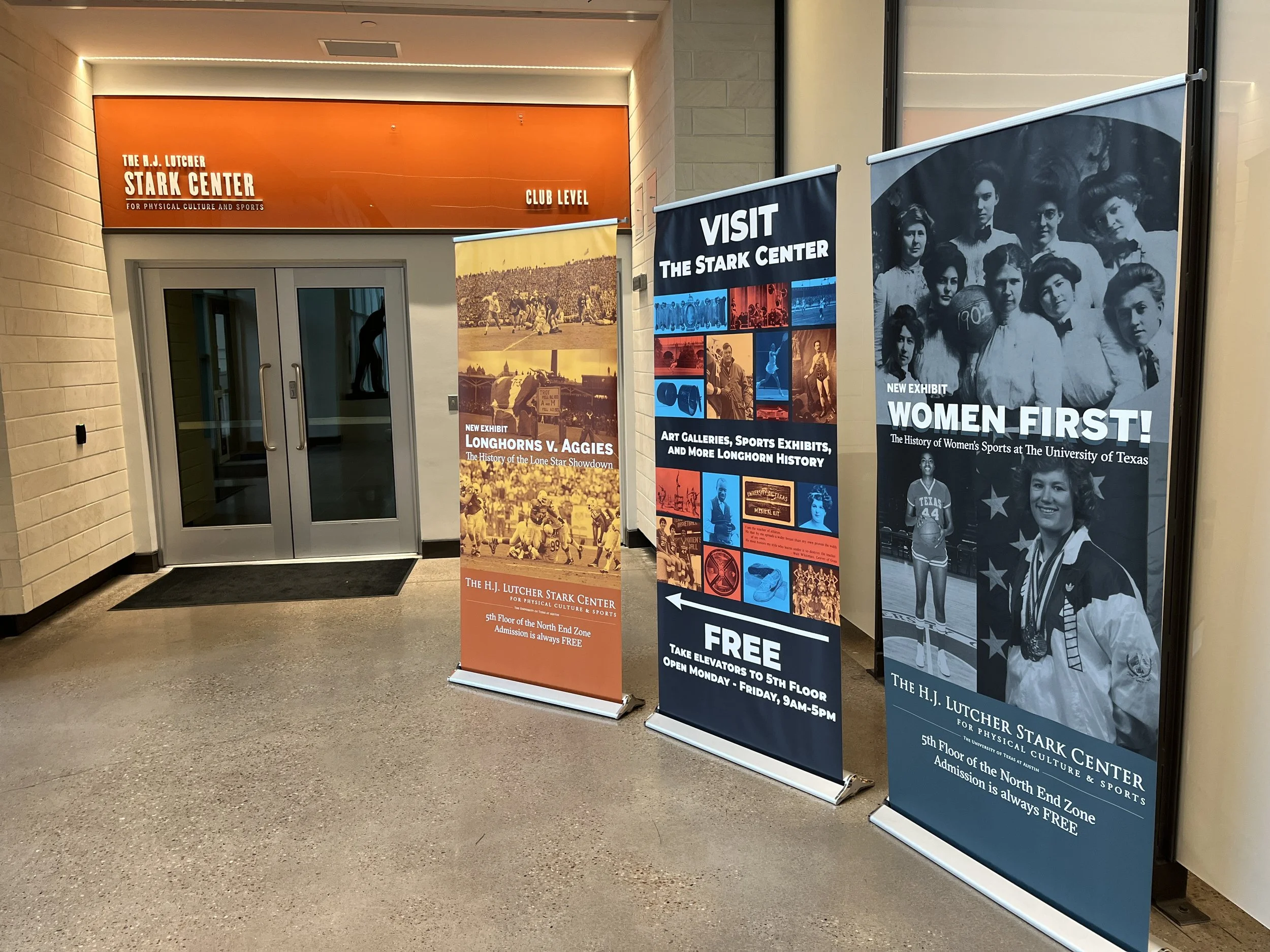 Entrance to the Stark Center for Physical Culture and Sports with three informational banners about current exhibits on women in sports, Longhorns vs. Aggies history, and general Stark Center exhibits. The entrance has double glass doors, and a sign 