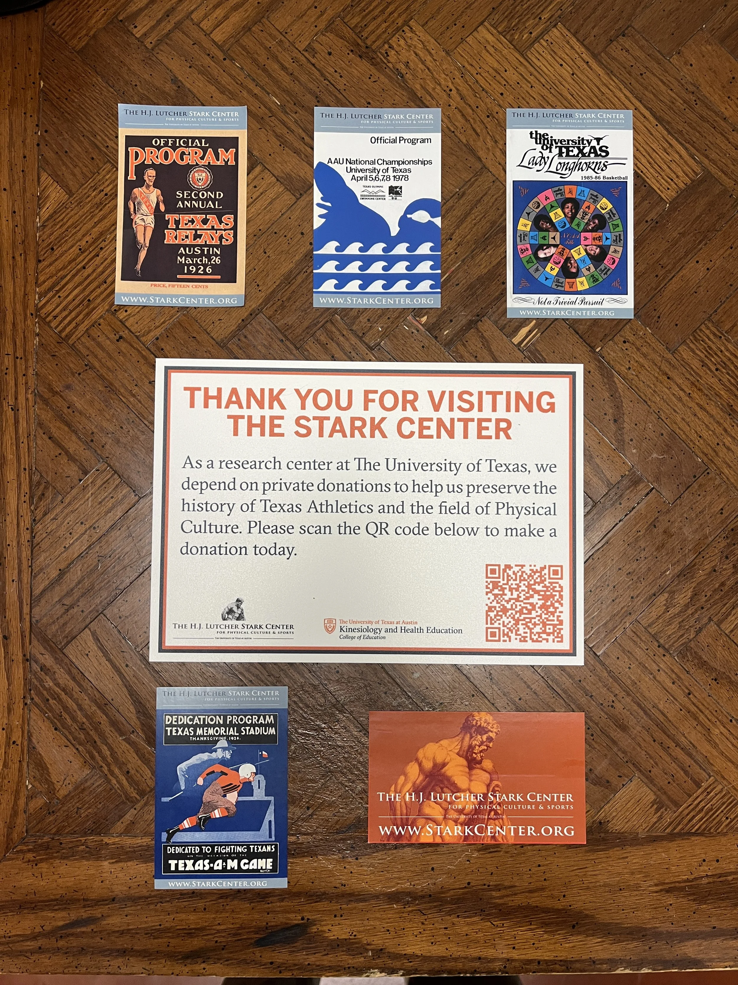 Collection of posters and a flyer on a wooden surface related to The University of Texas athletics and the Stark Center, including an official program for Texas Relays 1926, an AAU Championships program 1978, a Lady Longhorns basketball poster, a tha