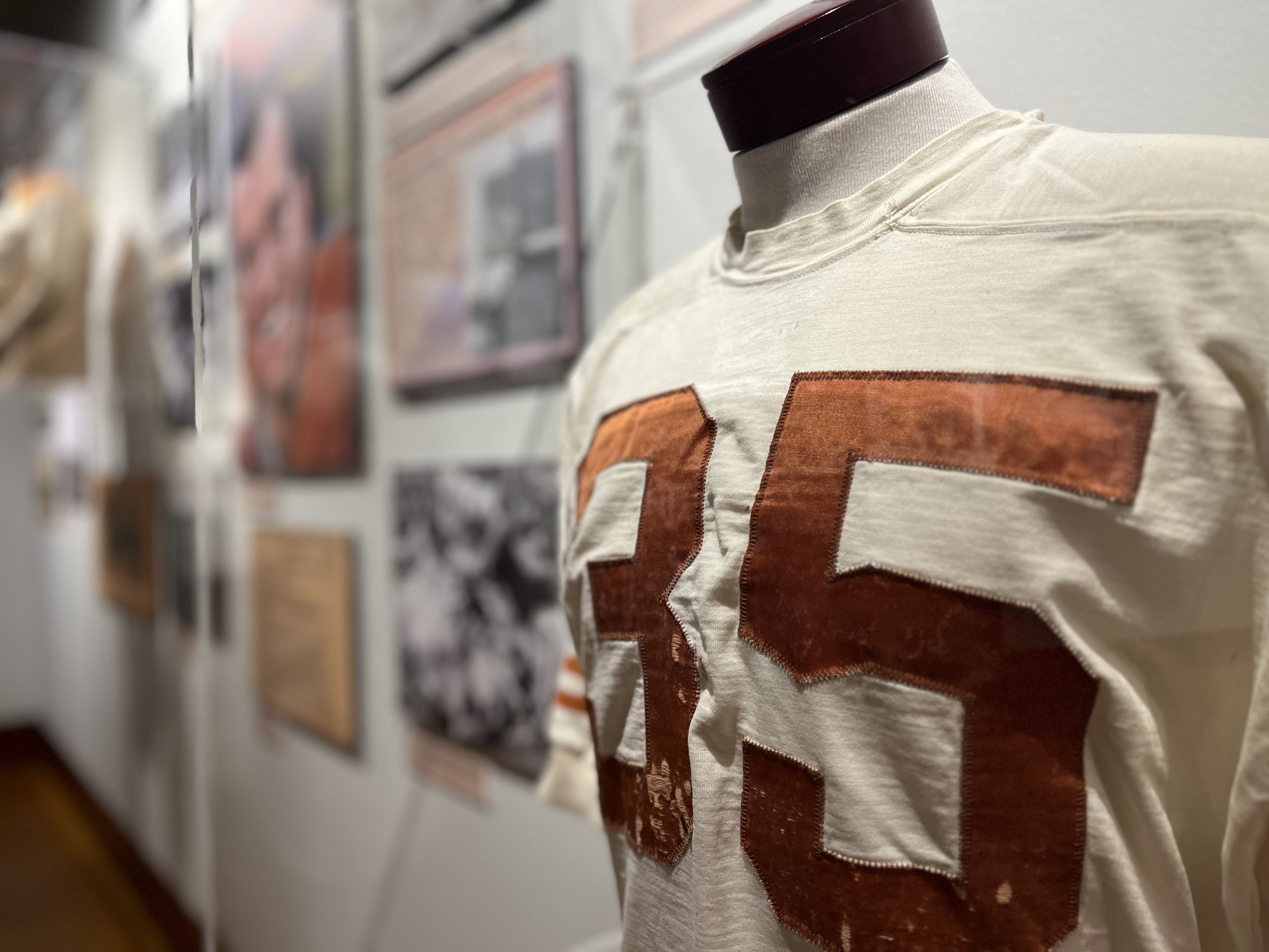 A mannequin wearing a white T-shirt with a large burnt-orange number 35 on the front, displayed in an art gallery or exhibit with photographs on the wall in the background.