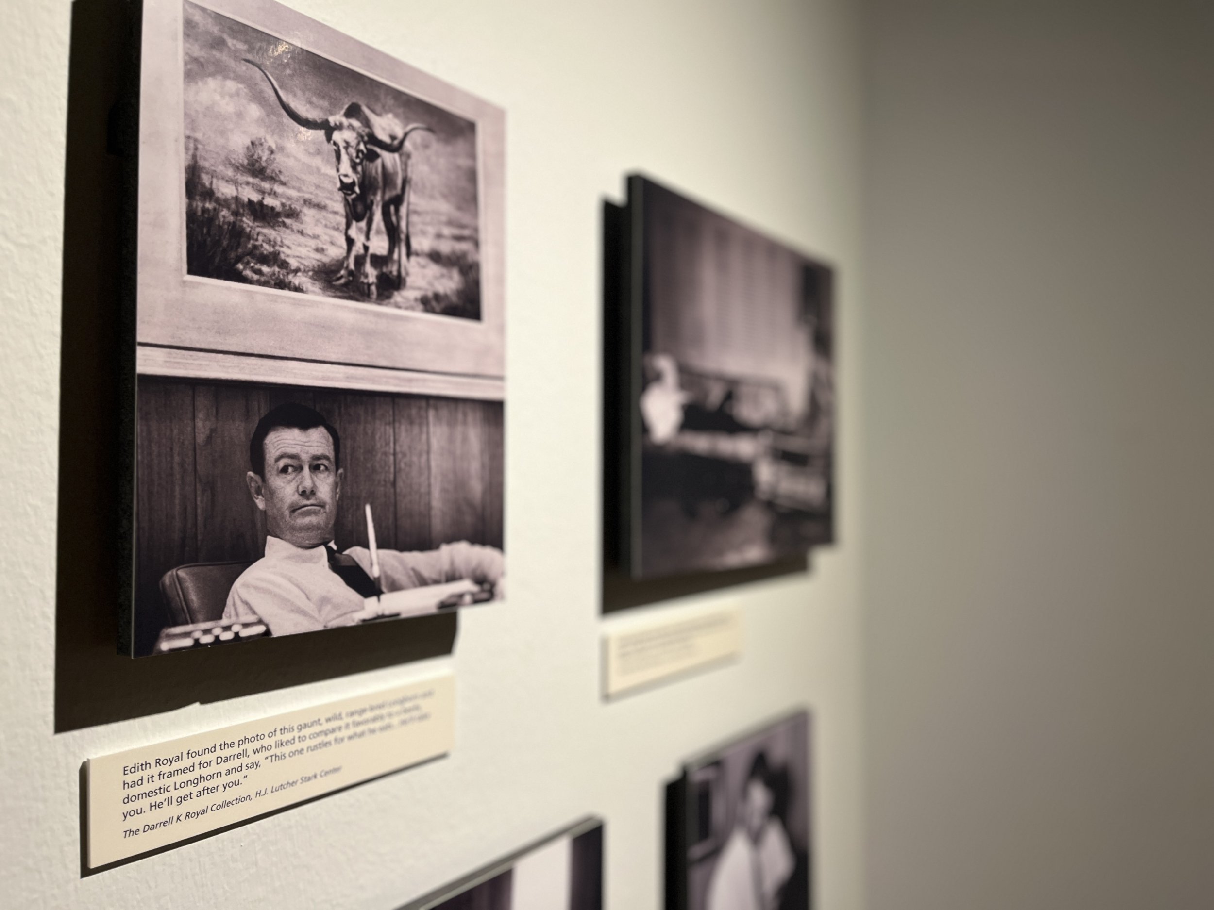 Black and white photo of a man sitting at a desk with a pen in his hand, looking to the side, framed above a painting of a longhorn steer.
