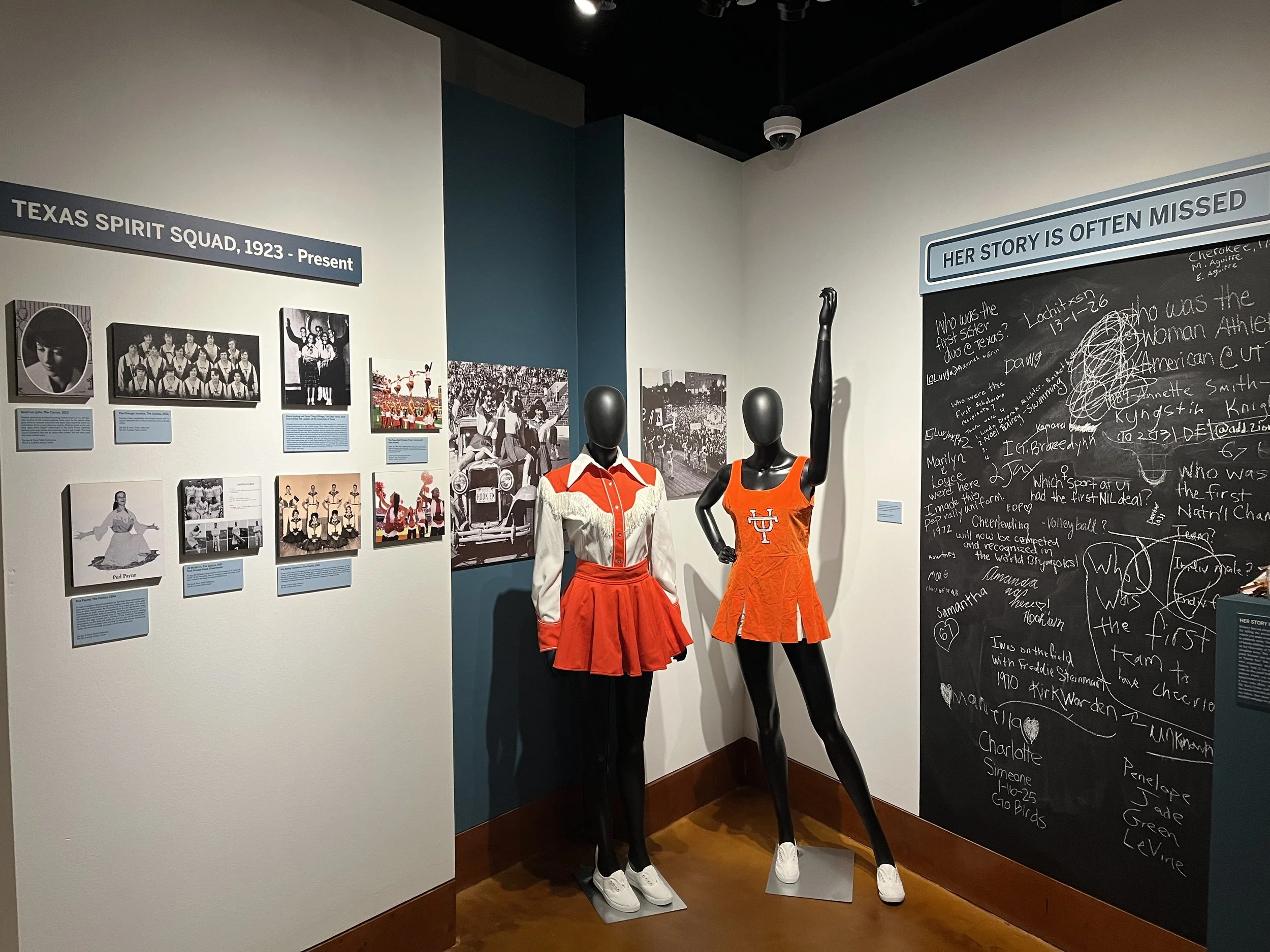 Exhibit of Texas Spirit Squad uniforms from 1923 to present, including two mannequins dressed in cheerleading outfits, one with a long sleeve shirt and another with a sleeveless dress, next to a wall with photographs, black and white images, and hand