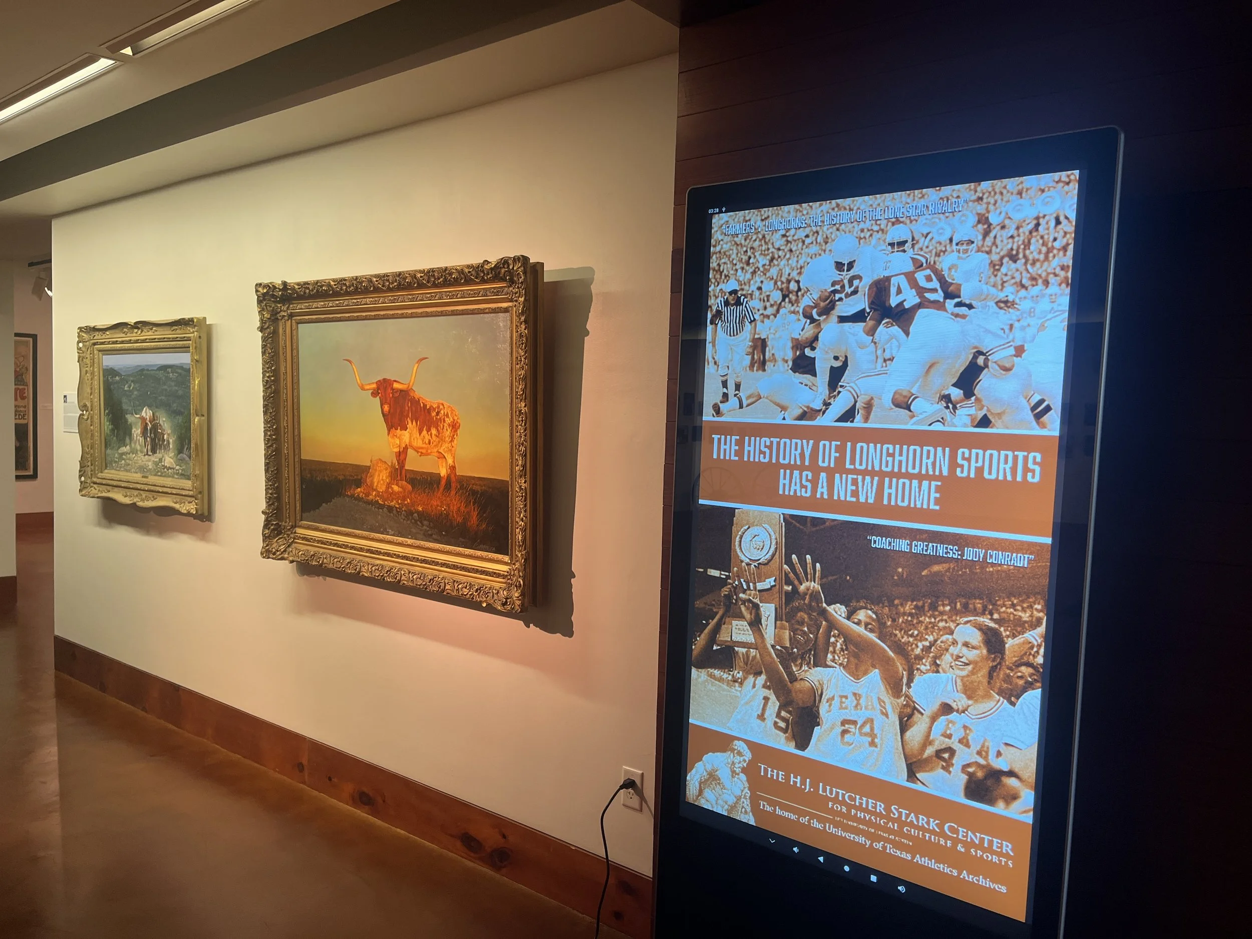 Digital display board in an art gallery showing the cover of a publication titled "The History of Longhorn Sports Has a New Home" with images of football players and a woman celebrating with a trophy, related to the University of Texas athletics.