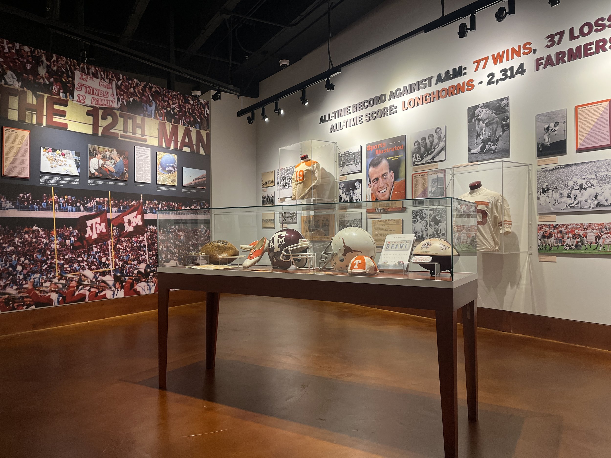 Display case containing football helmets and memorabilia, with photographs and information about Texas and Texas A&M football history on the wall behind.