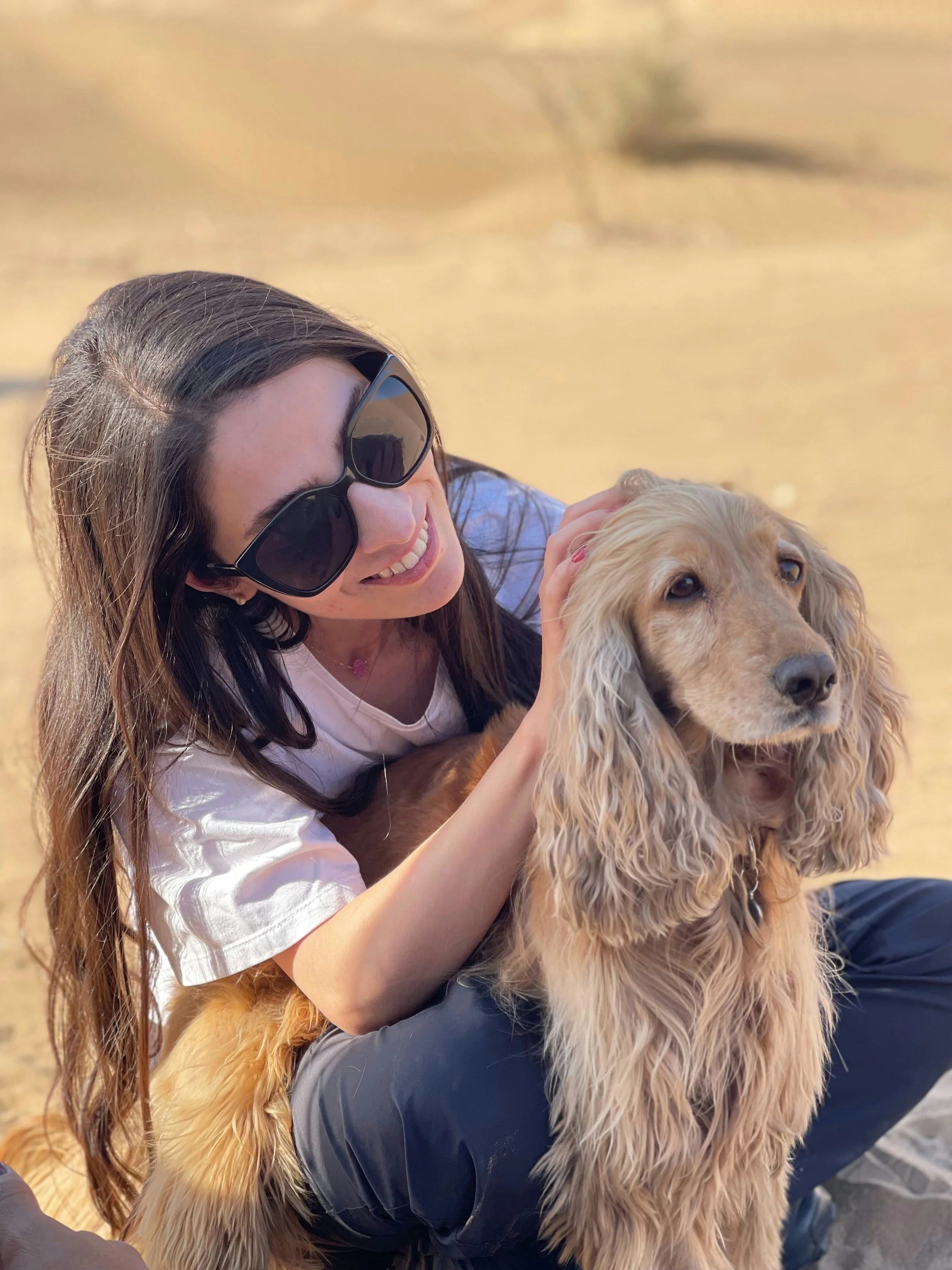 A woman with long brown hair wearing large sunglasses, smiling, sitting on the ground outdoors, petting a tan cocker spaniel.