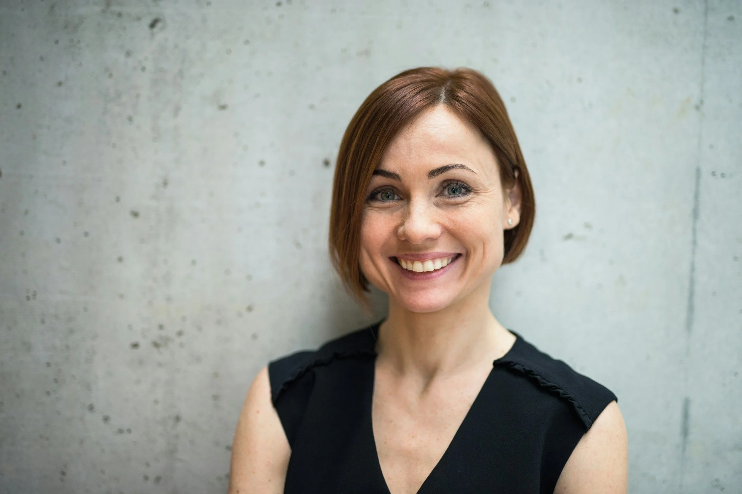 Close-up of a smiling woman with short red hair, wearing a black sleeveless top, standing against a concrete wall.