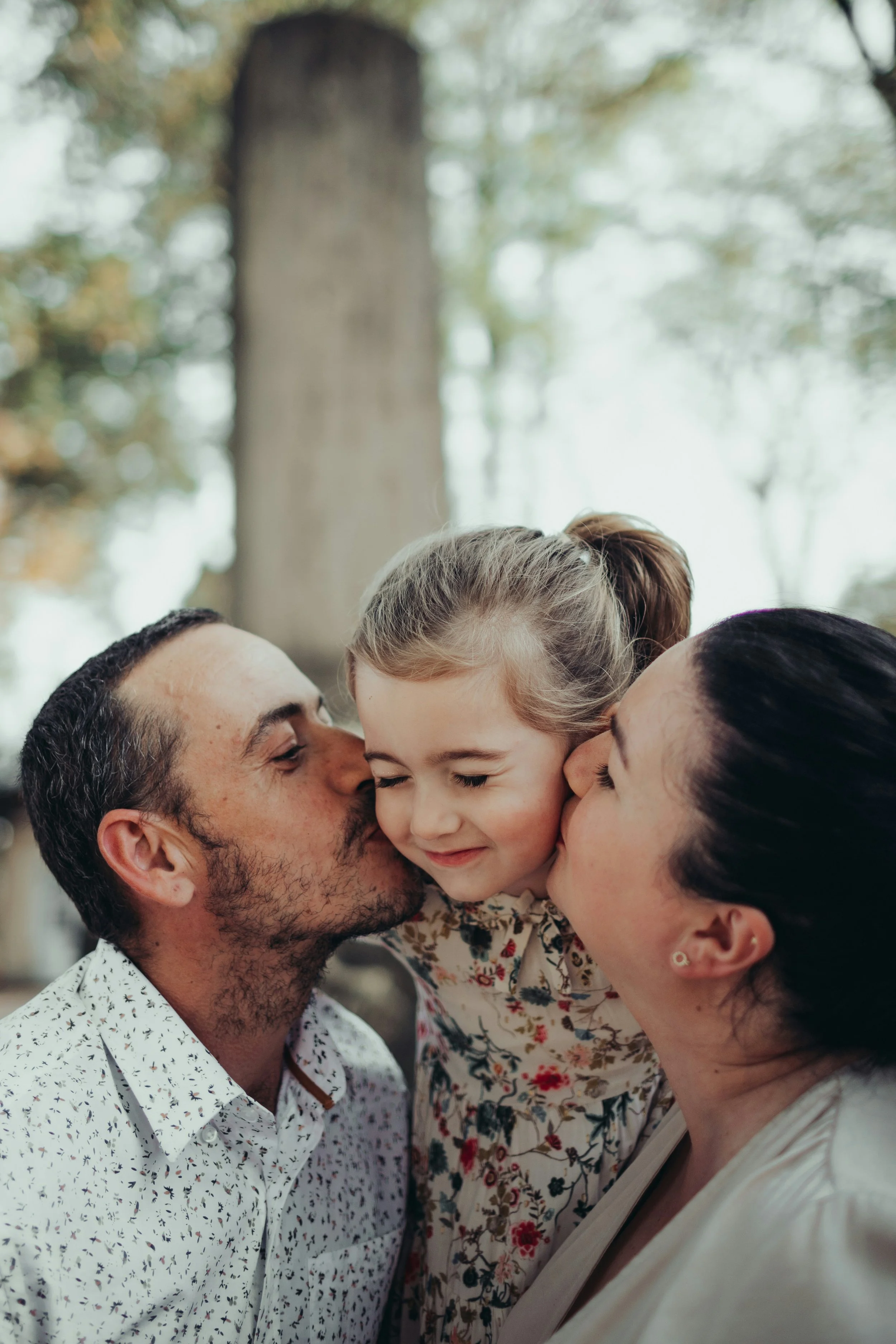 A man and woman kissing their young daughter on the cheeks outdoors, with trees and a large tree trunk in the background in Scotts Valley.