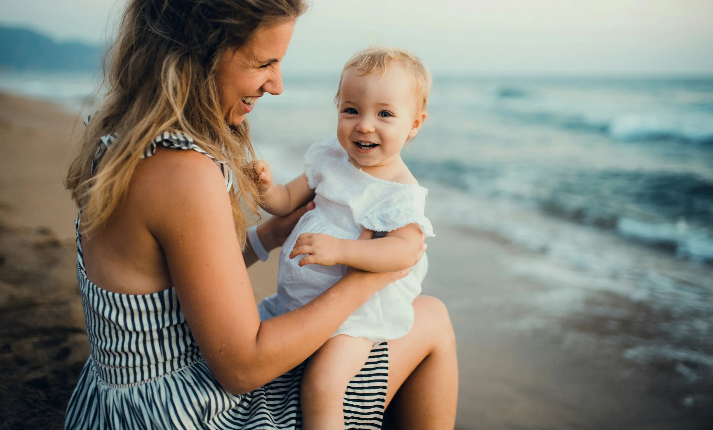 Laughing mother holding her baby on a beach — gentle pediatric chiropractic care for infants and children at Rhythm Health Santa Cruz