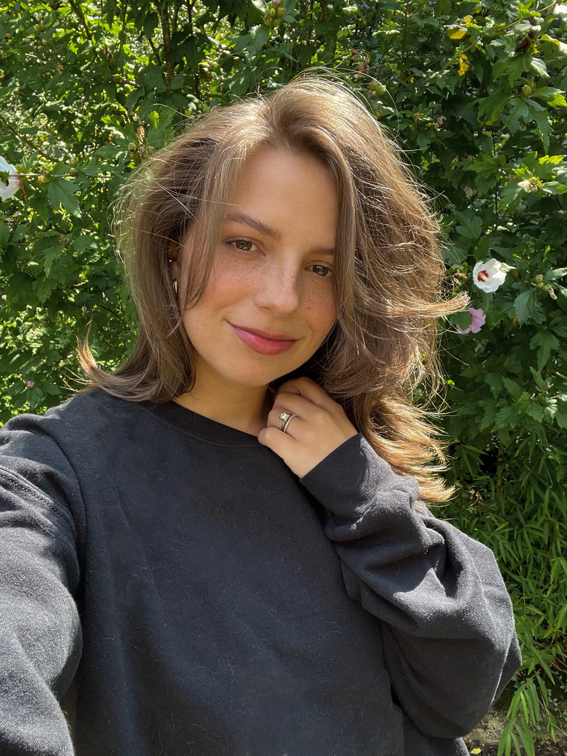 A woman with shoulder-length brown hair, freckles, and wearing a black sweatshirt is standing outdoors in front of green leafy plants with some white and pink flowers. She is smiling, touching her neck with her right hand, and wearing a ring on her ring finger.