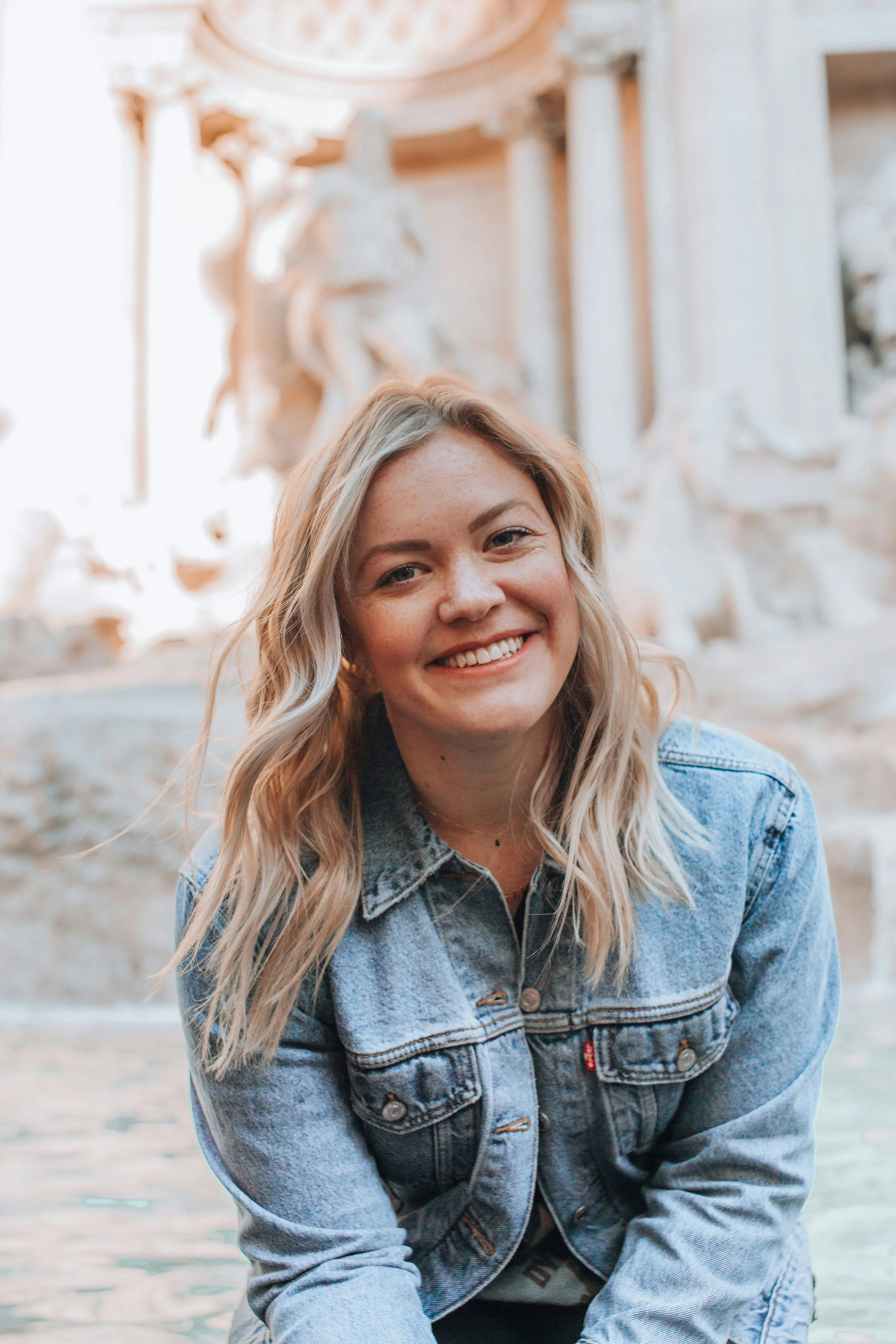 A smiling woman with blond hair in a denim jacket sitting near water with a classical sculpture and architecture in the background.