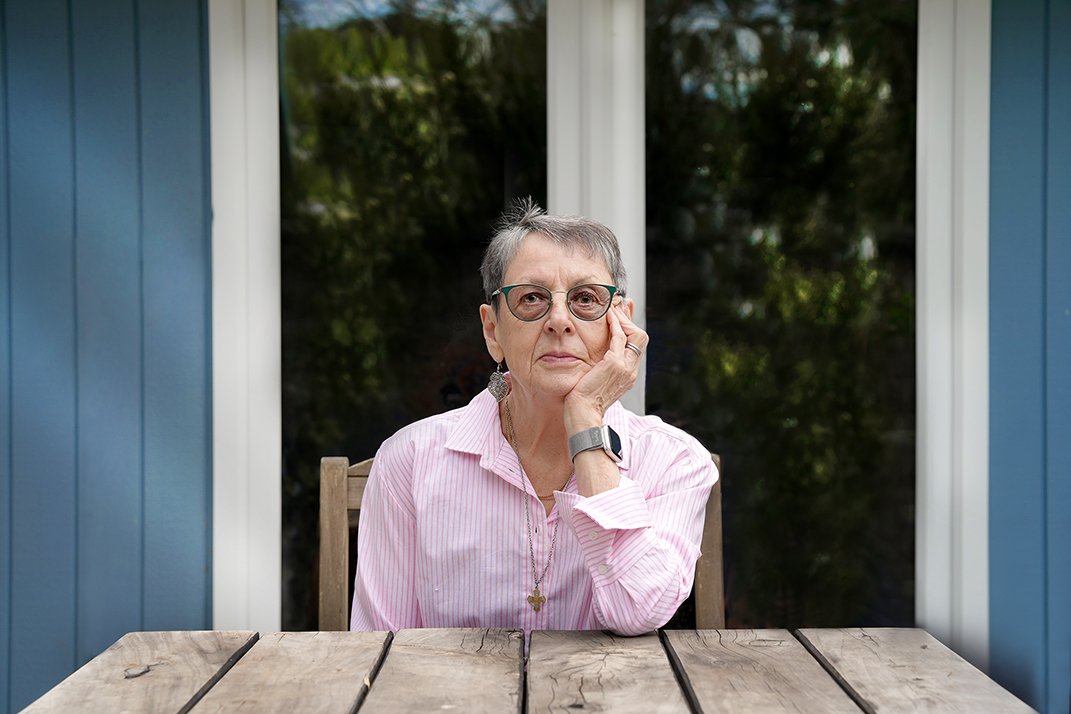 Older woman with short gray hair, glasses, and a pink striped shirt sitting at a wooden table outside her house, resting her head on her hand and looking at the camera.