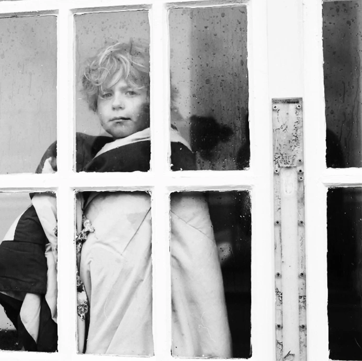 Child with curly hair looking through a front door window, which is divided into multiple panes, with a face showing a somber or contemplative expression.