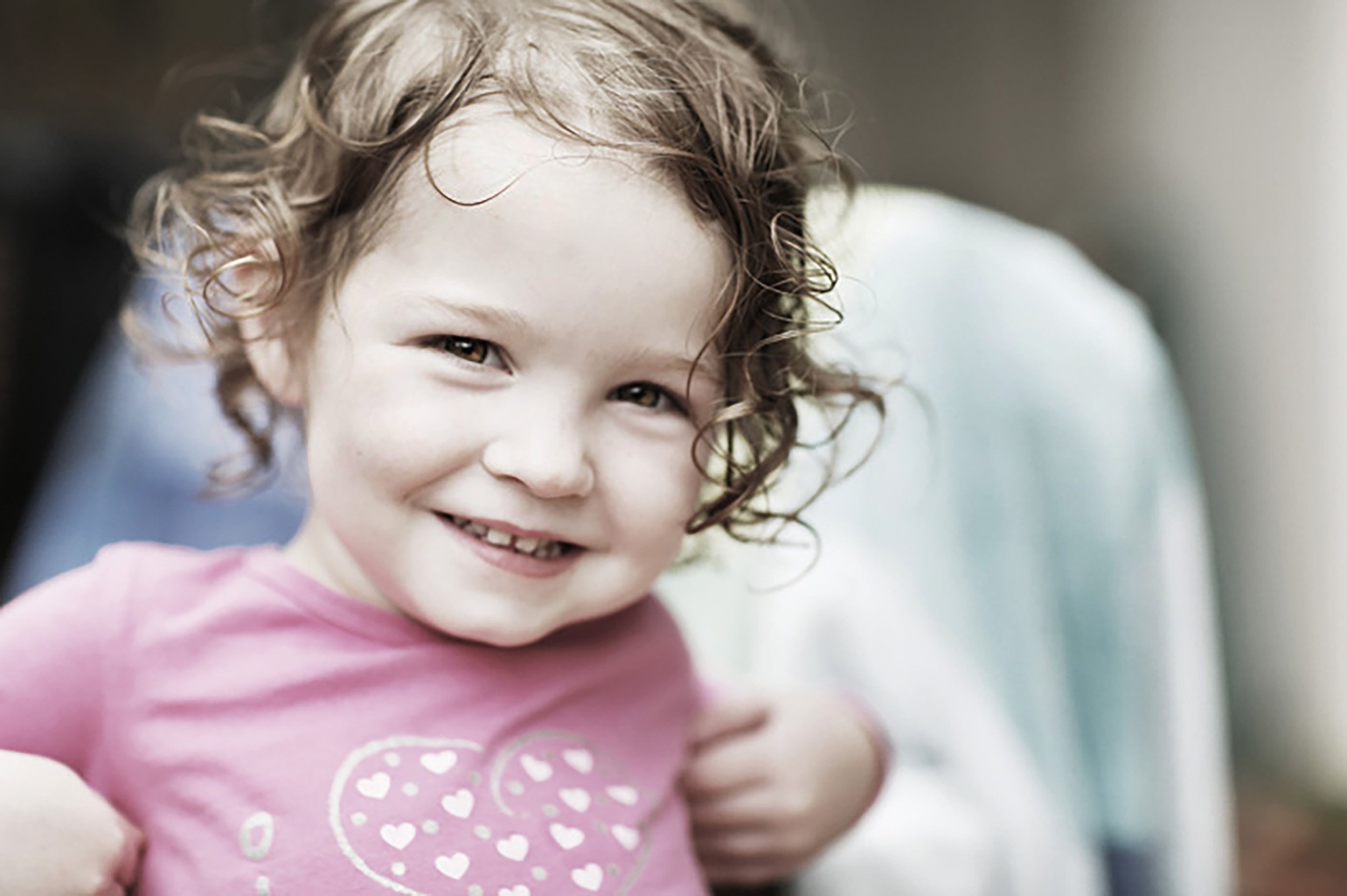 Young girl with curly hair smiling at camera, wearing a pink shirt with a heart design.