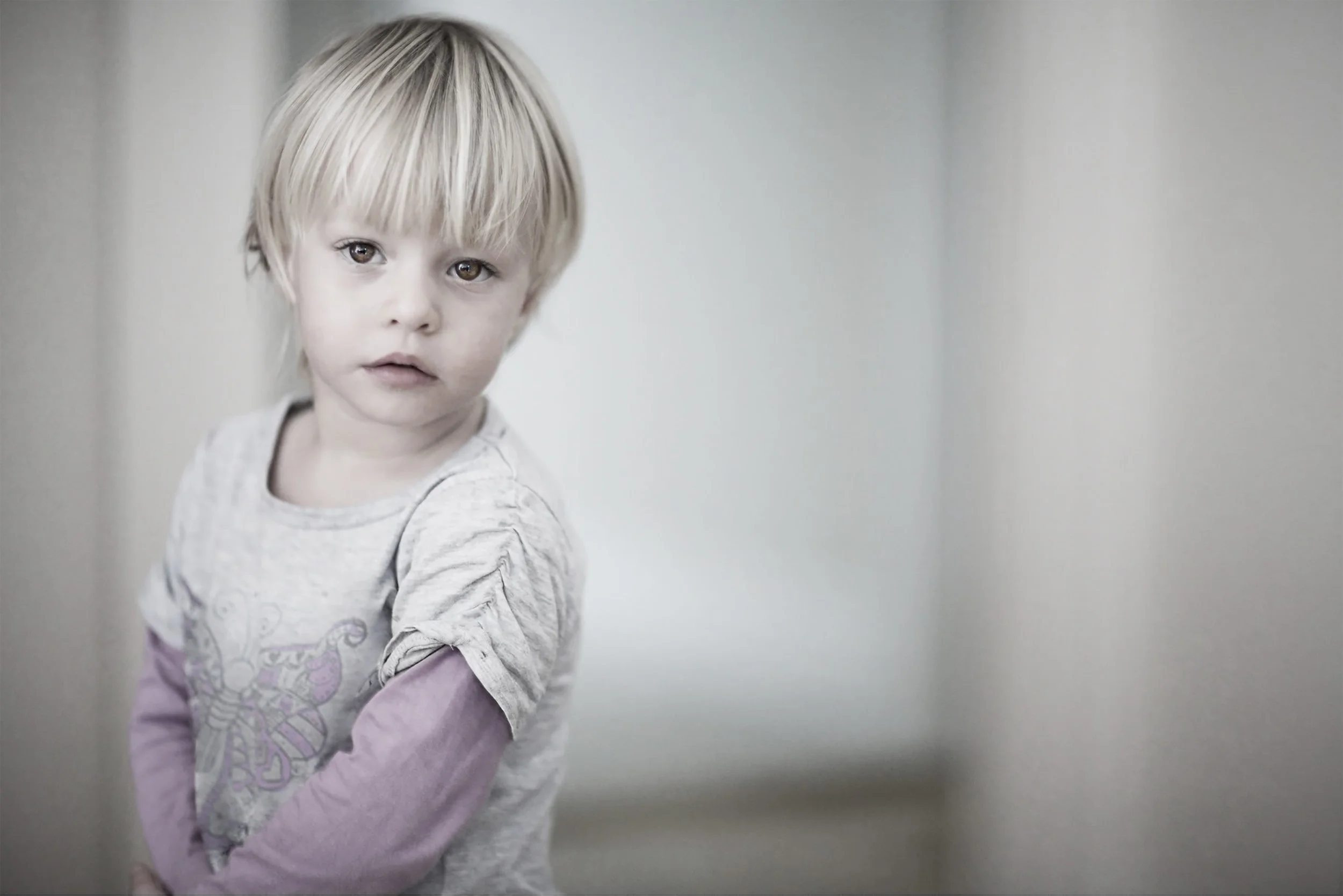 A young blonde girl with a serious expression, wearing a light gray and pink long-sleeve shirt, standing indoors against a plain light-colored wall.