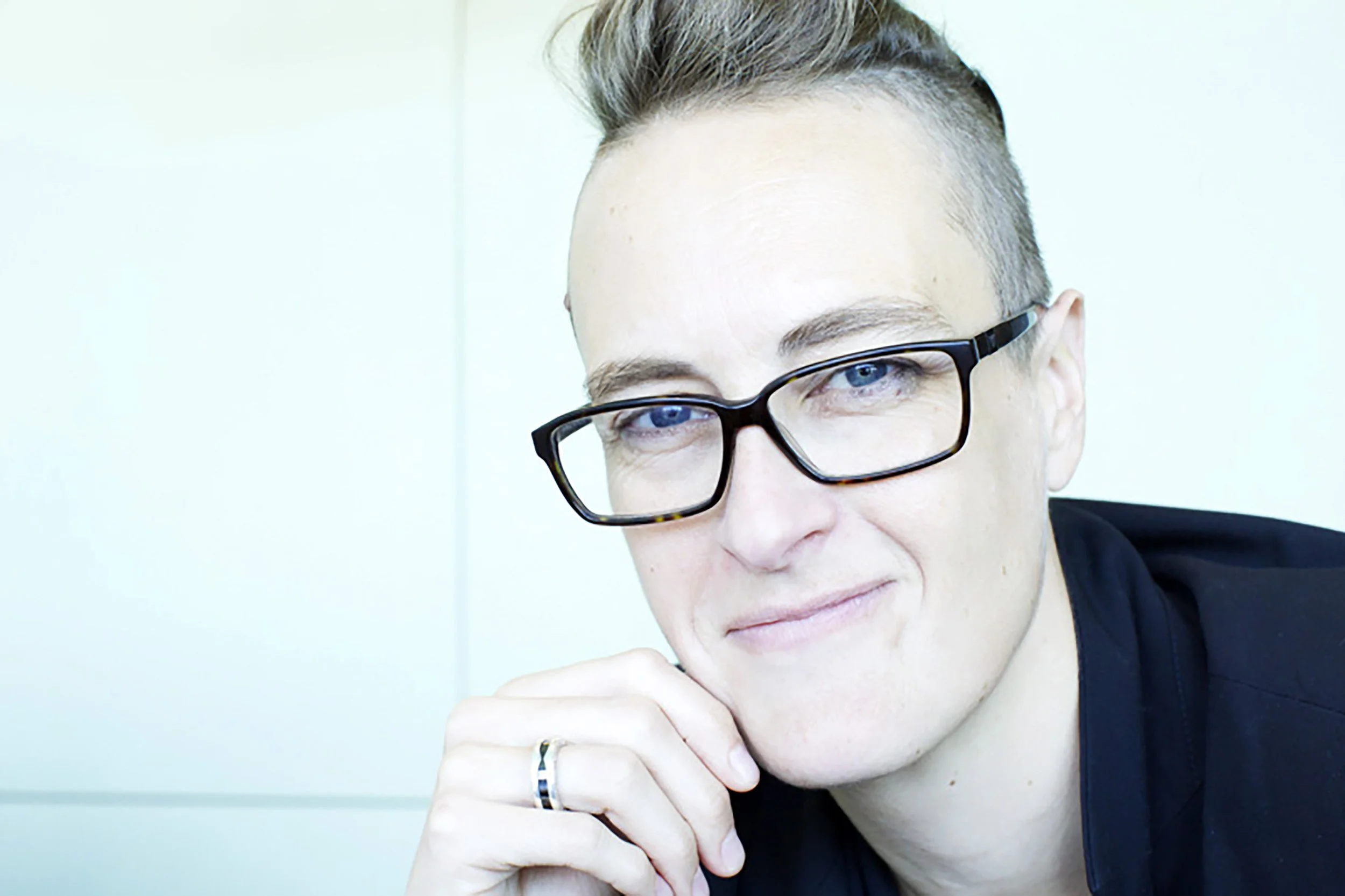 A young man with light skin and short, styled hair wearing black glasses and a black shirt, resting his chin on his hand and looking at the camera with a slight smile.