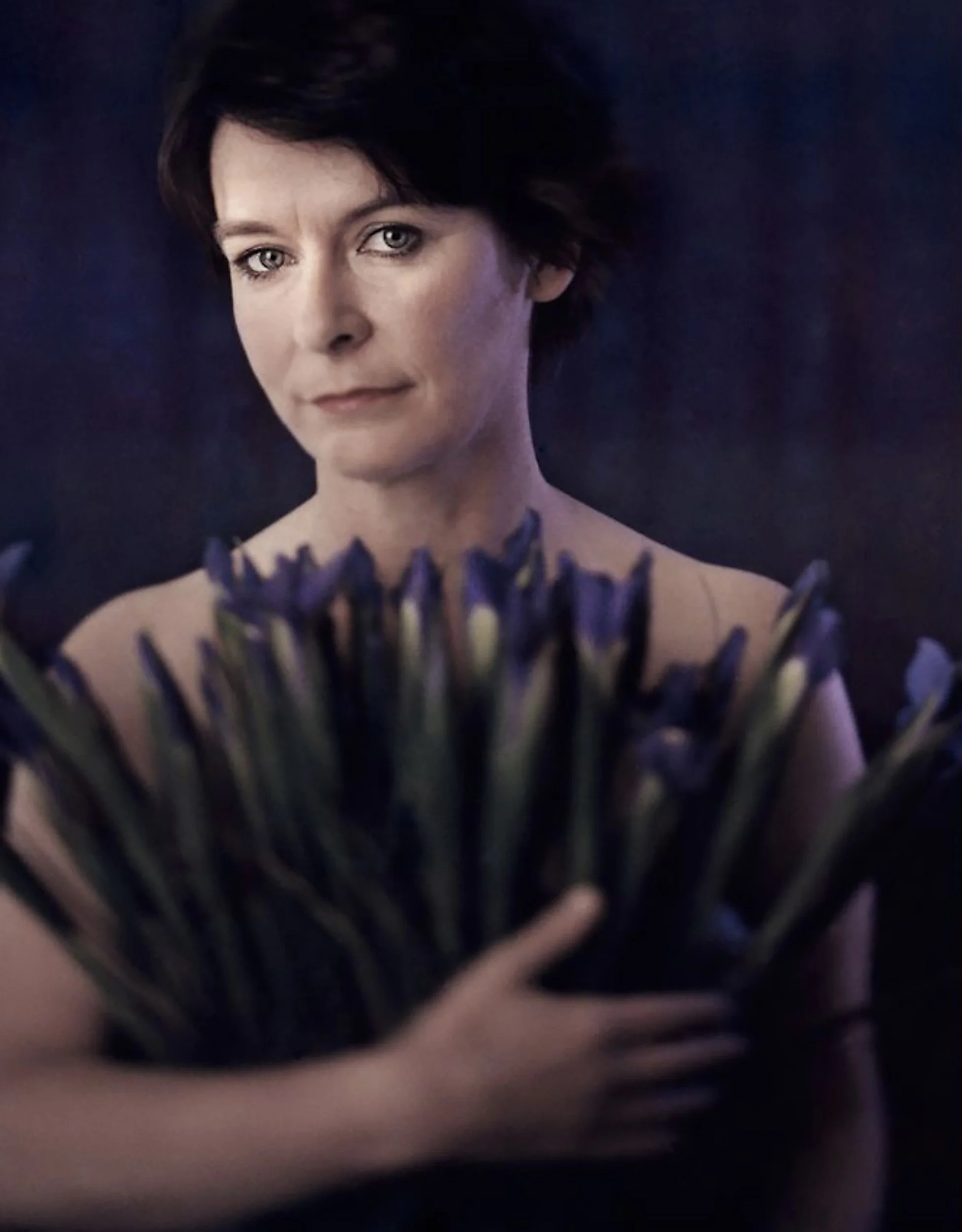 A woman with short dark hair holding a large bouquet of purple irises, looking directly at the camera with a serious expression, against a dark background.