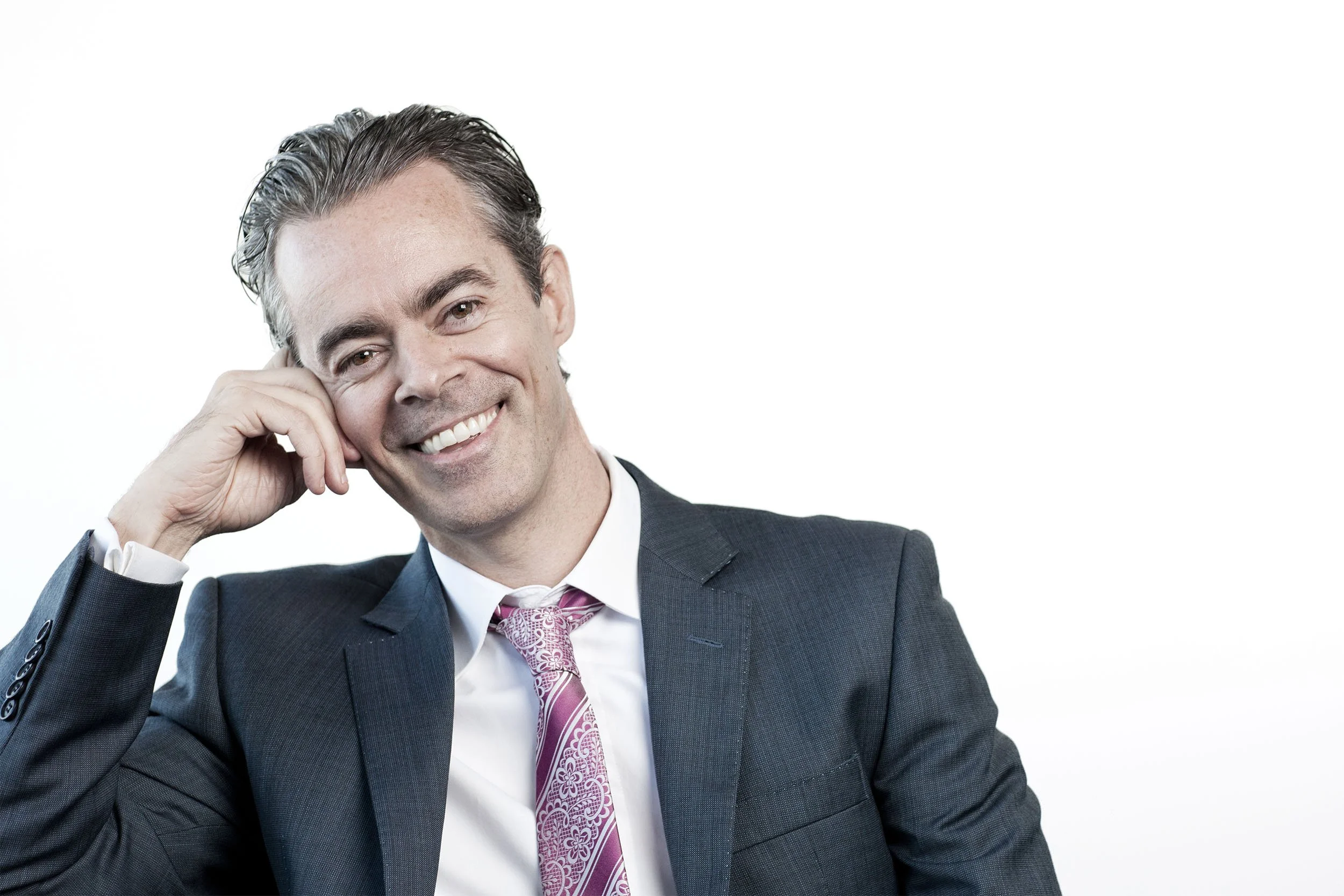 A smiling professional man in a dark suit and pink patterned tie, leaning on his hand, in a relaxed pose against a white background.