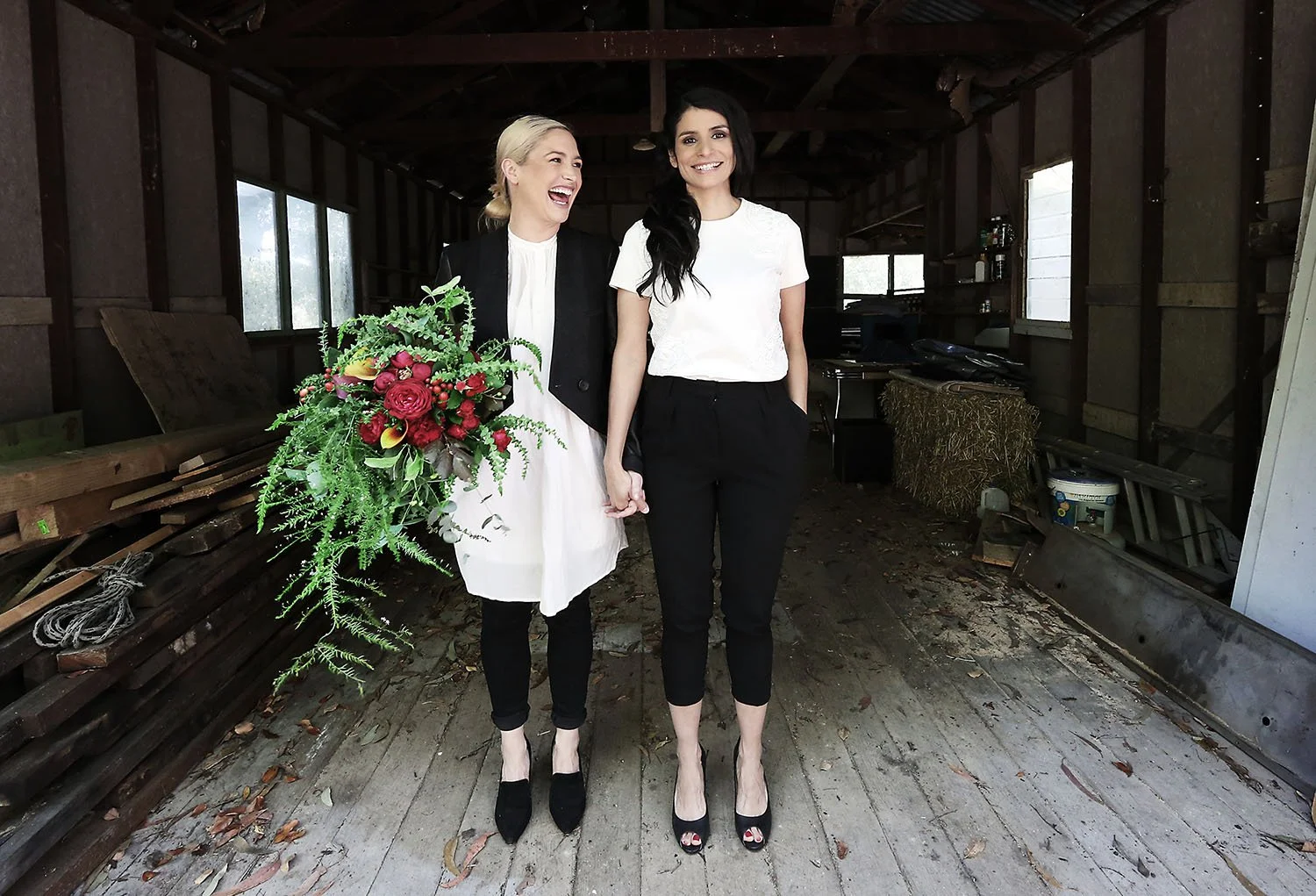 Two women are standing inside a rustic, partially open barn. One woman, with blonde hair, is smiling and holding a large bouquet of red and pink flowers. She is wearing a white dress with a black jacket. The other woman, with dark hair, is smiling an