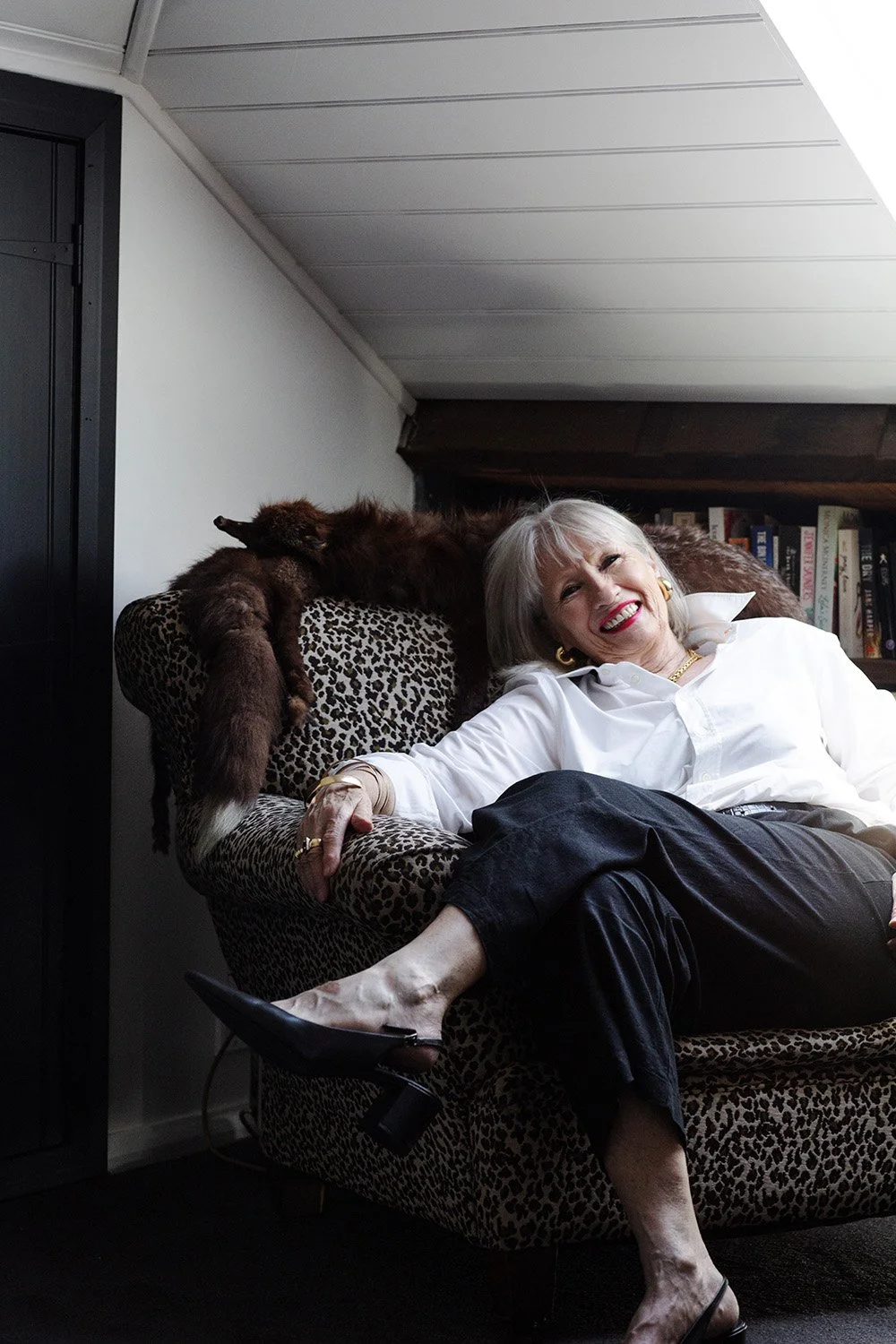 A joyful elderly woman with blonde hair and a white blouse sitting on a leopard print armchair, smiling at the camera. A small brown dog is lying on the backrest of the armchair, stretching out with its head and front legs hanging over the side.