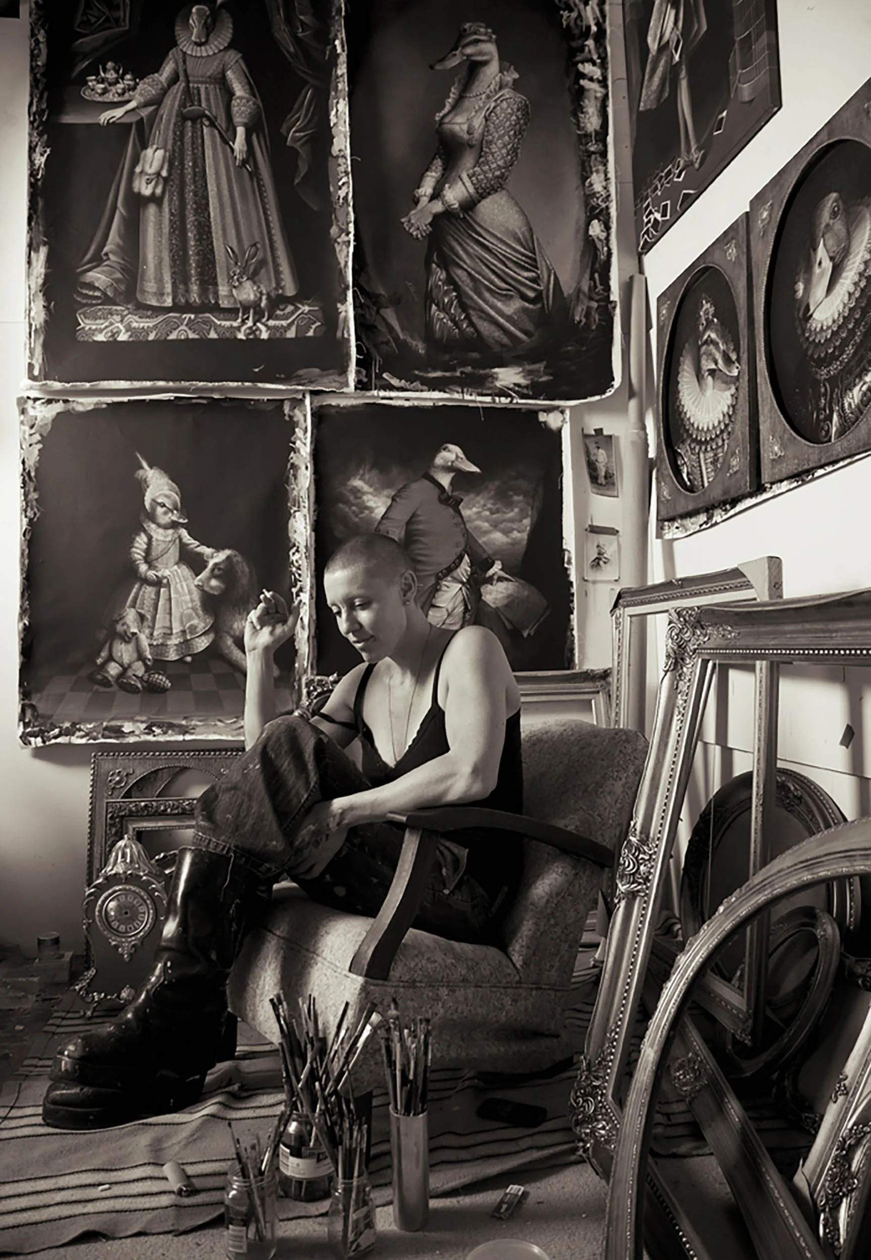 A woman sitting in an art studio with various large, framed fantasy and portrait paintings hanging on the walls, and art supplies like paintbrushes and framed artworks scattered around.