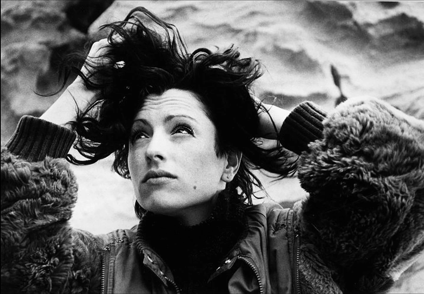 A black-and-white photograph of a woman with short, curly hair, wearing a warm fur-lined coat, lying outside with her hands in her hair and looking up.