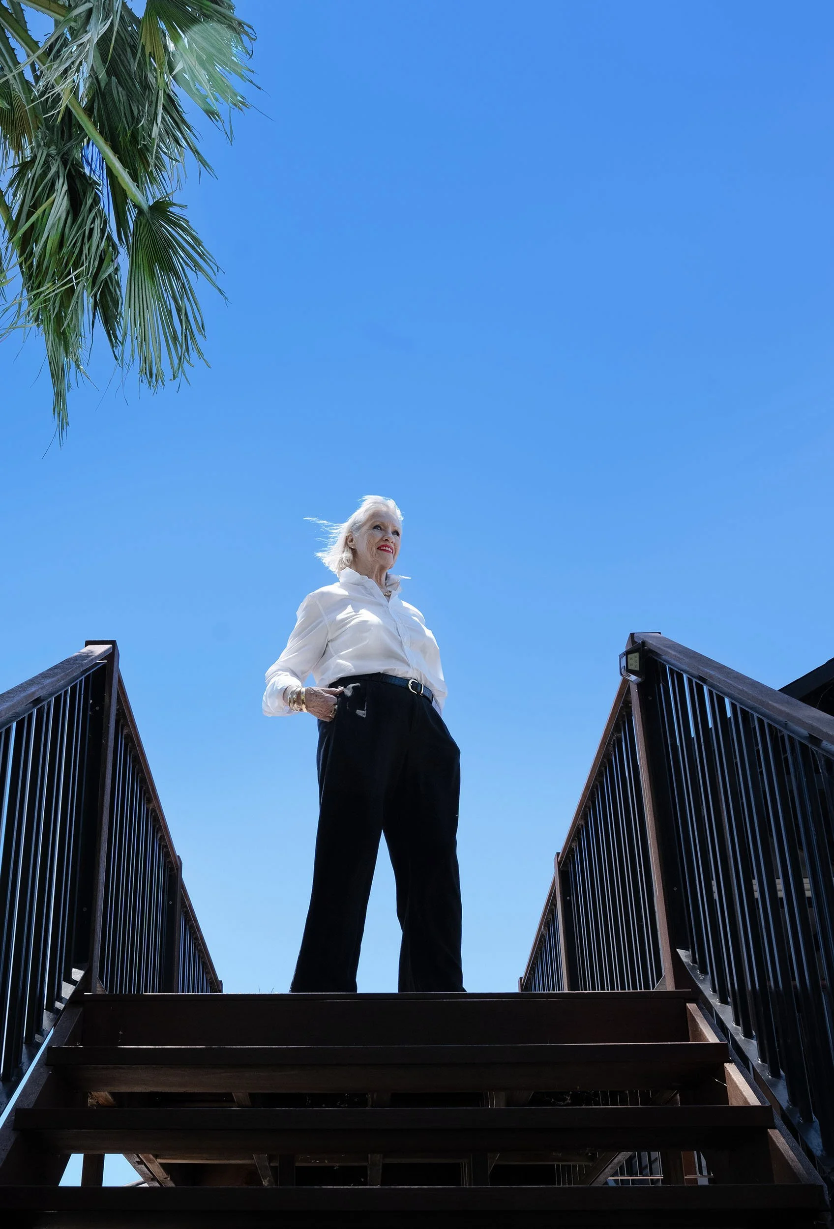 An elderly woman with white hair standing on a wooden staircase outdoors under a bright blue sky, with green palm leaves visible in the top left corner.