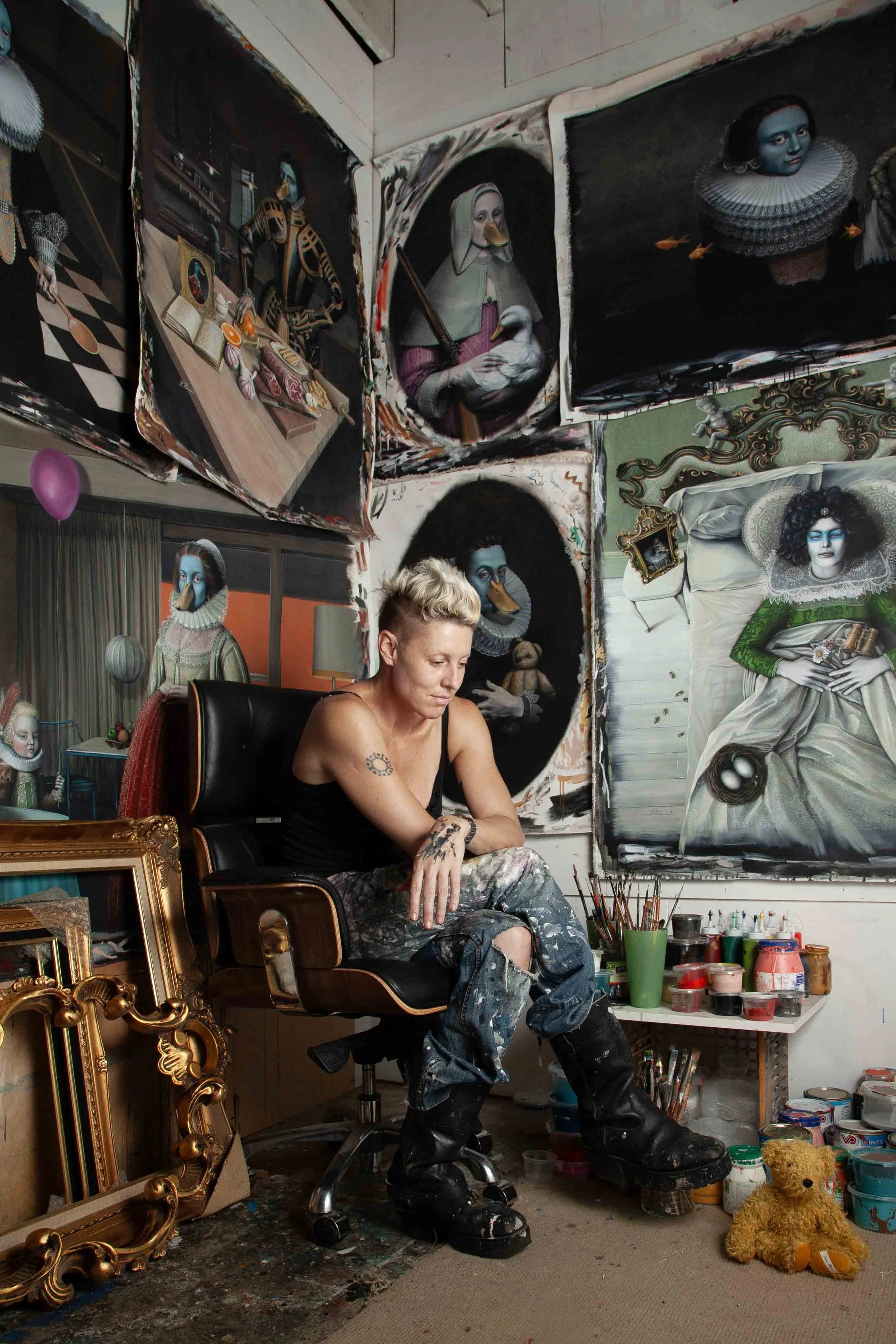 An artist with short blonde hair and tattoos sitting in a black chair in her studio, surrounded by colorful and surreal portrait artworks of people with duck-like features on the walls.